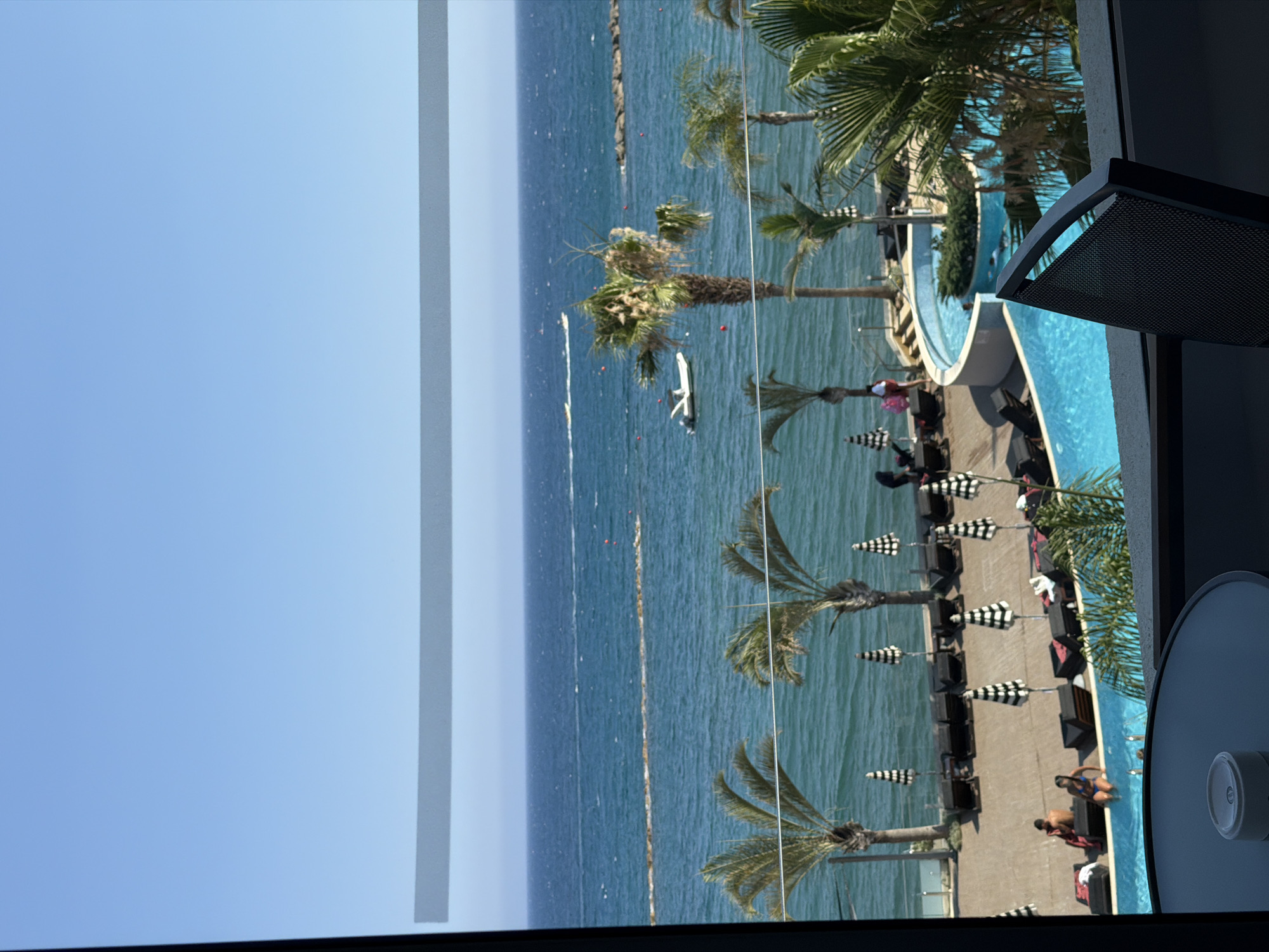 This media file depicts a vibrant outdoor scene with a focus on leisure activities. The primary subjects are people enjoying themselves near and in a resort-style swimming pool, adjacent to a body of water that appears to be the sea. Palm trees are scattered throughout the scene, suggesting a tropical or Mediterranean location.
The activity taking place is clearly relaxation and recreation. People are seen lounging by the pool, some are in the water, and others are walking. The presence of lounge chairs, umbrellas with a checkered pattern, and a well-maintained pool area points towards a resort or a high-end vacation spot. There are also a few boats visible in the water, indicating the possibility of water sports or transportation.
Notable details include the bright, sunny weather, suggested by the clear blue sky and the strong sunlight casting shadows. The time of day appears to be daytime, likely midday or afternoon, given the intensity of the light. The water in the pool is a striking turquoise blue, contrasting with the deeper blue of the sea. The overall atmosphere seems relaxed and cheerful.
The image is framed by what appears to be a window or a balcony railing, with some dark structural elements visible on the right side. There are also some vertical gray bars on the left side of the image, possibly indicating digital interference or a compositional choice. No visible text is present in the media file.