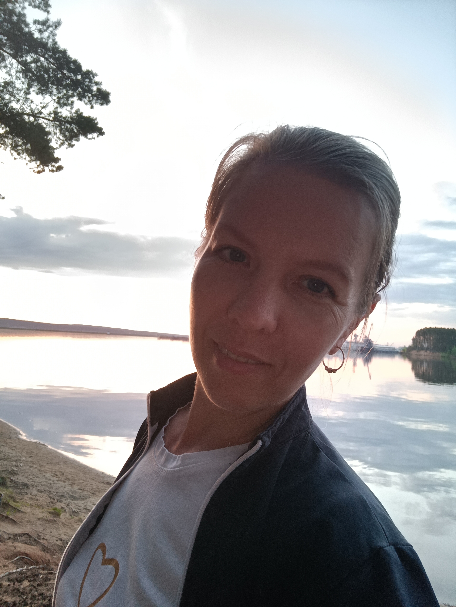 A selfie of a woman with short grey hair, wearing a white t-shirt with a gold heart and a dark blue zip-up jacket, is taken outdoors near a body of water. The woman is fair-skinned and smiling. The background shows a lake with reflections of the sky and trees. On the far side of the lake, there are buildings and trees, possibly indicating an urban area. To the left, there's a beach with light-colored sand and vegetation. A tree branch is visible in the upper left corner of the image. Given the location specified as Chaikovsky, Russia, it can be assumed that the lake is the Kama Reservoir.