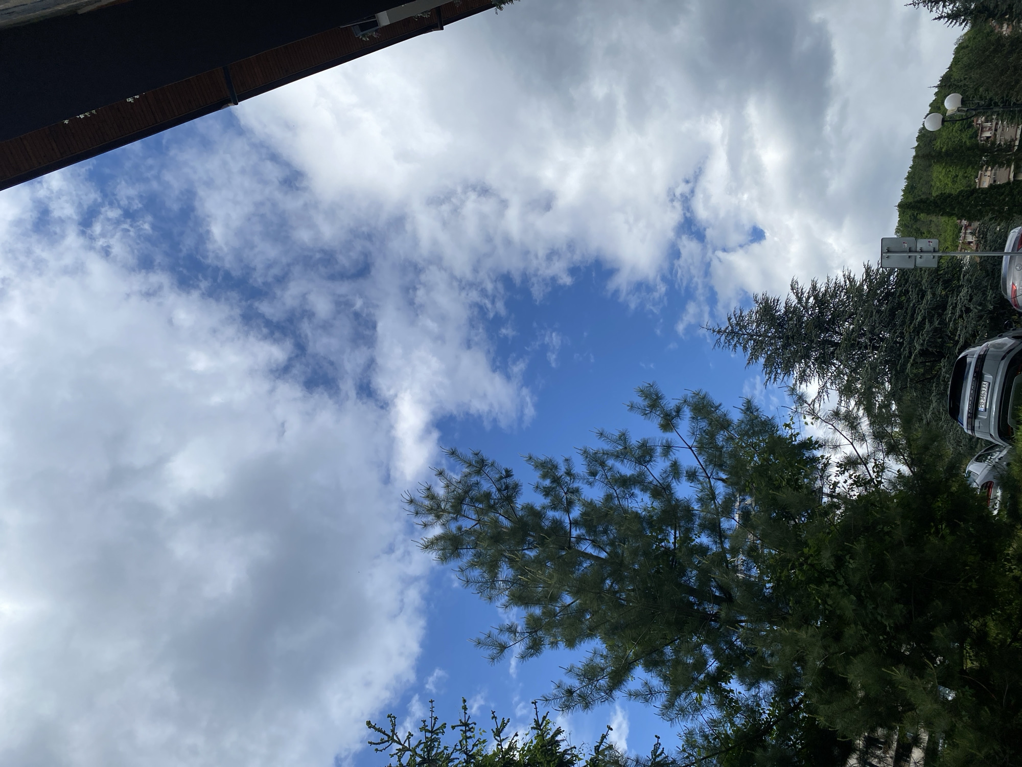 The image is a low-angle shot taken outdoors. A portion of a building is visible at the top left. The rest of the scene is dominated by a sky filled with white clouds against a blue sky. Several trees are visible at the bottom and right of the frame. Cars are parked to the right with their registration plates visible. A road sign is also partially visible amongst the trees. The photo was likely taken in or around the city of Smolyan, Bulgaria.