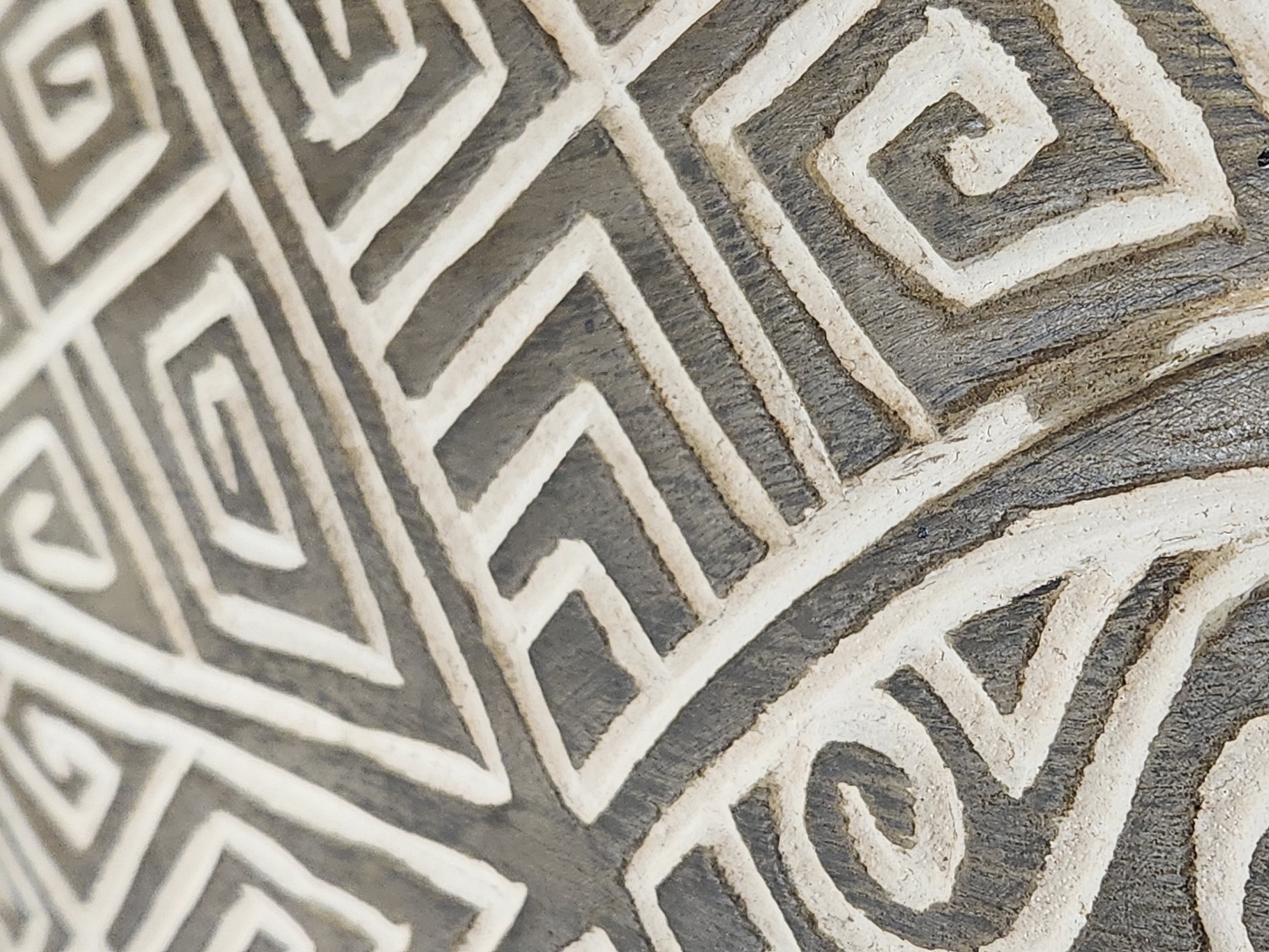 The image is a close-up, high-angle shot of a textured surface with a repeating geometric pattern. The pattern consists of interlocking squares and spirals, rendered in contrasting light beige and dark gray colors. The texture appears to be carved or incised into the material, with visible striations and some roughness. The lighting is diffused, creating soft shadows that emphasize the depth of the carved elements. There are no people, animals, or specific landmarks visible in the image. The scene is abstract and focuses solely on the intricate design and tactile qualities of the surface. The exact location in Maitland, United States of America, is not discernible from the visual information provided.