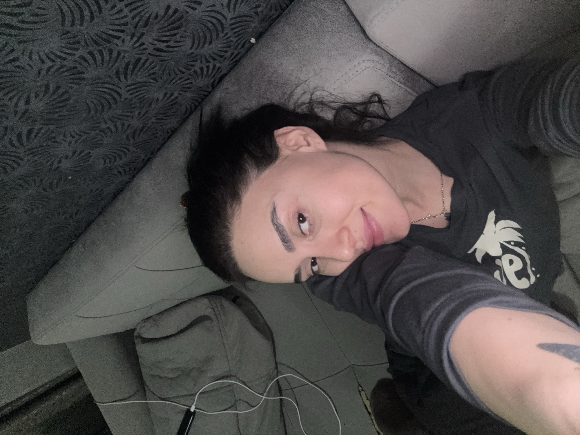 A person is lying on a grey couch in an indoor environment. The person has dark hair, fair skin, and is wearing a dark-colored shirt with a light-colored design. A part of their arm is visible, showing a tattoo. The couch is made of a soft material and has a pattern on the back. A white headphone cable is visible near the bottom of the frame. It is not possible to determine the location of this image in Tbilisi, Georgia from the given information.