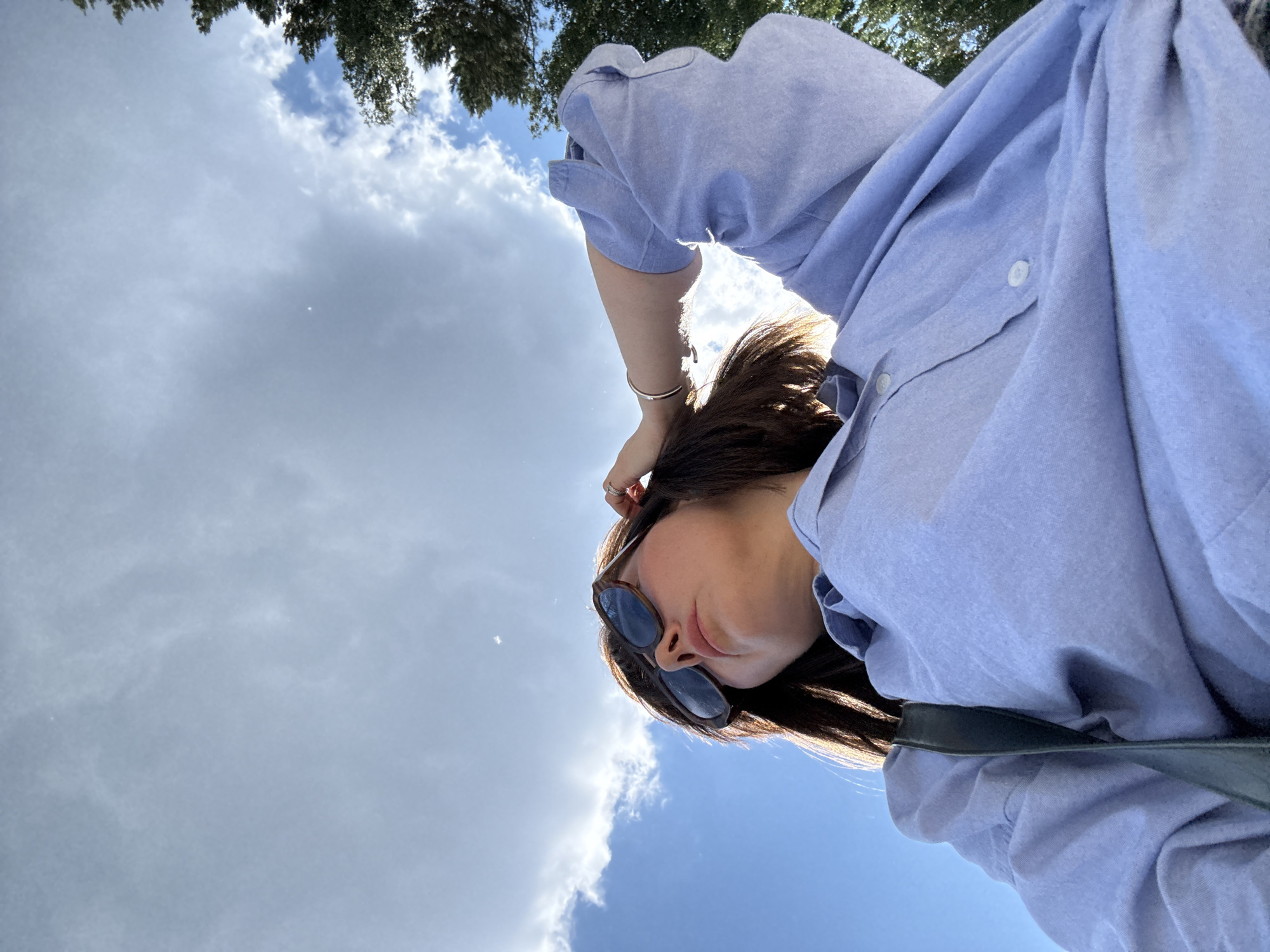 A low-angle, close-up, point-of-view shot shows a person wearing a light blue button-down shirt and sunglasses. The person's left arm is raised, with their hand resting on their head, and a silver bracelet is visible on their wrist. The background consists of a bright blue sky with scattered clouds, and the tops of green trees are visible on the right side of the frame. The lighting suggests it is daytime. The image was taken outdoors.