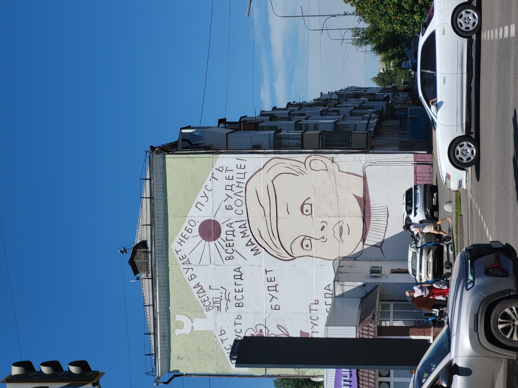 A mural depicting a child's face and various text phrases is painted on the side of a light-colored, multi-story apartment building. The mural includes phrases in Russian such as "ВСЕГДА БУДЕТ НЕБО, ПУСТЬ" and "ВСЕГДА БУДЕТ МАМА! СОЛНЦЕ!". A stylized depiction of a house with a bird on top is also visible. The building has balconies and windows. In the foreground, a silver SUV is in the left lane of a paved road. A white sedan is in the right lane. Several people, some with strollers and electric scooters, are standing on the sidewalk near the building. A traffic light is visible on the left side of the image. The sky is clear and blue, and trees are present in the background. The scene appears to be an urban environment in Petropavl, Kazakhstan.