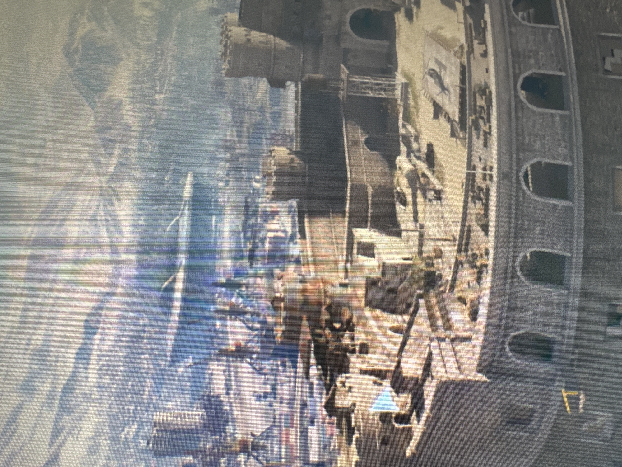 The image depicts a video game scene, likely from a first-person shooter, showcasing a large, fortified structure resembling a castle or fortress. The architecture features stone walls, turrets, and arched doorways. Within the fortress, there are various military elements, including what appear to be mounted guns or anti-aircraft emplacements scattered across the rooftops. A small, light blue flag is visible on a battlement.
In the background, a city skyline is visible, characterized by modern high-rise buildings and industrial elements like cranes and docks. Further in the distance, snow-capped mountains and a large, oval-shaped structure, possibly a stadium or arena, are present. The overall environment suggests a cold, possibly wintery, setting. The scene does not contain any identifiable people. The city of Koltsovo, Russia, is not explicitly depicted or referenced within the visual information of the image itself.