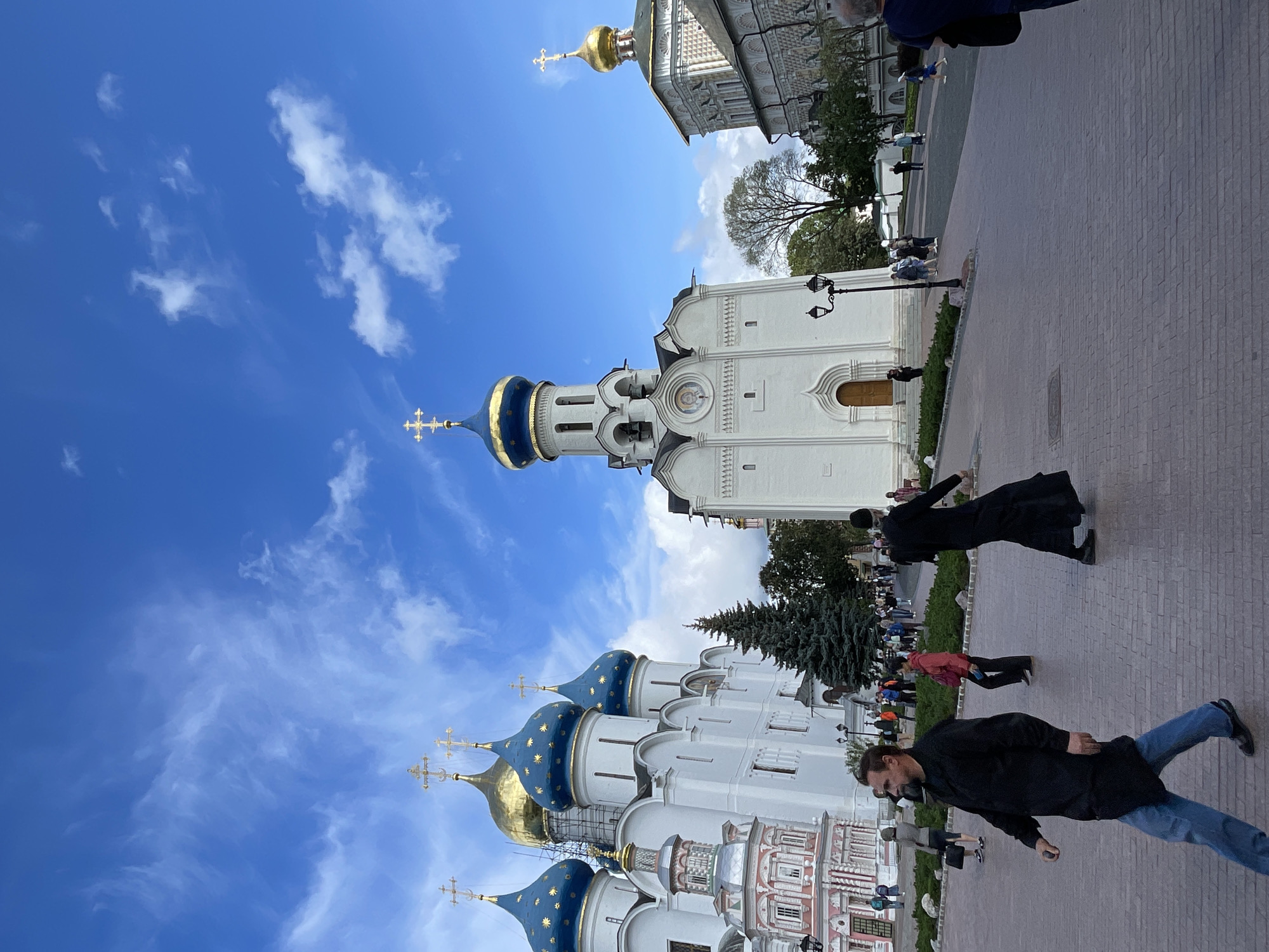 The image depicts a wide-angle view of a large, open courtyard in front of several ornate religious buildings, likely the Trinity Lavra of St. Sergius in Sergiyev Posad, Russia. Dominating the background are several white buildings with distinctive golden and blue-domed roofs topped with crosses. To the left, a cluster of these buildings features multiple blue domes with gold stars and a prominent gold dome. A smaller, pink and white tiered structure is attached to this complex. In the center, a taller, white bell tower with a blue and gold dome and a cross stands prominently. To the right, another white building with a gold dome and intricate decorative patterns is visible.
The courtyard is paved with brickwork. Several people are present in the scene. In the foreground, a man in a black, knee-length coat and blue jeans is walking towards the left, looking to his left. Further into the courtyard, another person, also in black attire that appears to be a robe, is walking towards the right. In the middle ground, a crowd of people, appearing to be tourists and locals, are dispersed throughout the courtyard, some walking, others gathered in small groups. A few individuals are seen in the background near the buildings. Evergreen trees are scattered in the middle ground, with more trees visible further in the distance. The sky above is a vibrant blue with scattered white clouds. A lamppost stands in the mid-ground near the bell tower.