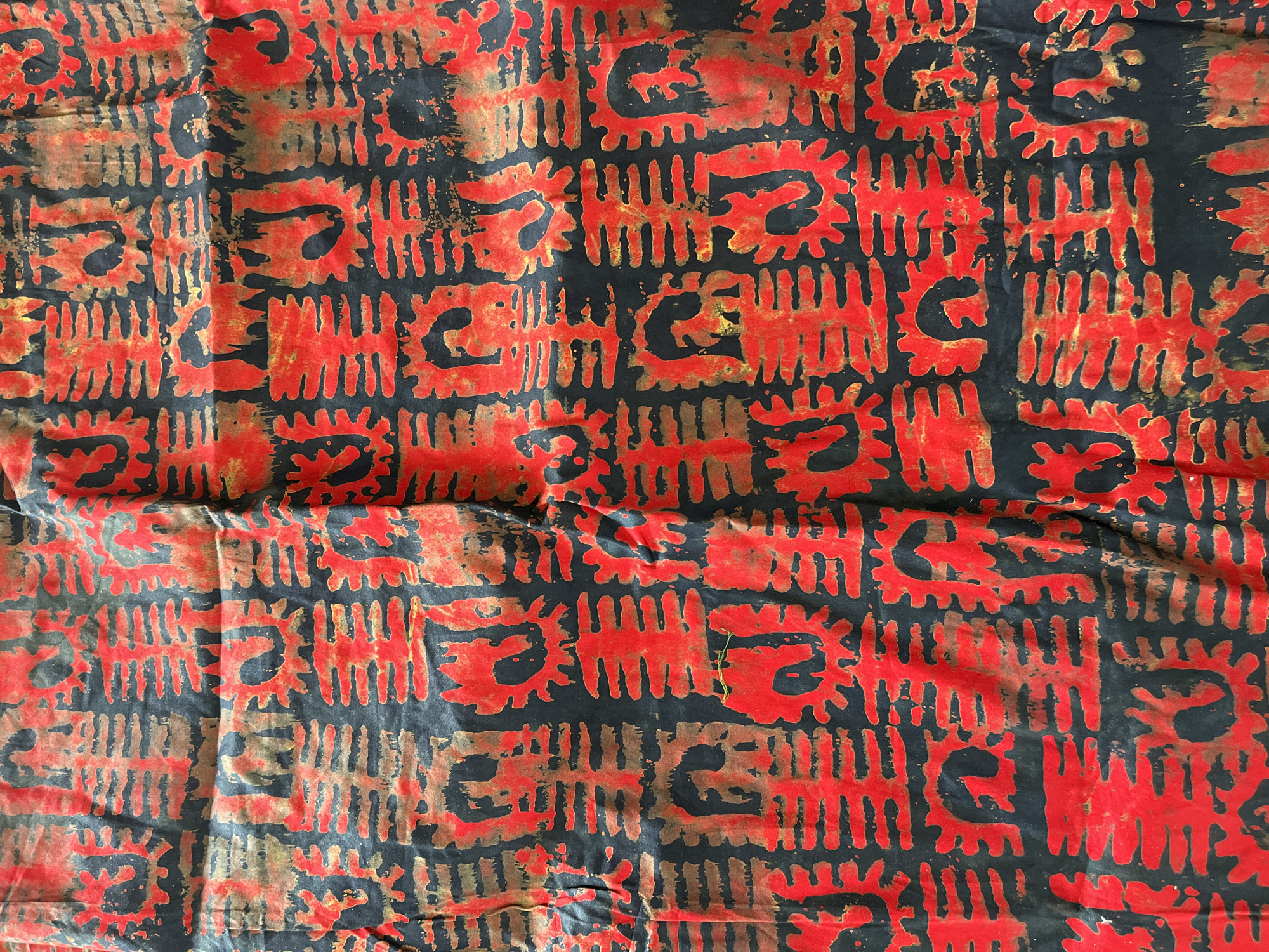 The image shows a piece of patterned textile, likely a fabric swatch. The primary colors are red and black, with some areas showing a bronze or brown undertone. The pattern appears to be a repeating motif consisting of short vertical lines interspersed with stylized, abstract shapes reminiscent of symbols or figures. The textile is not perfectly flat and shows some folds and creases. There is no information about Jos, Nigeria or any other location. No people or actions are visible. The environment is limited to the fabric itself.