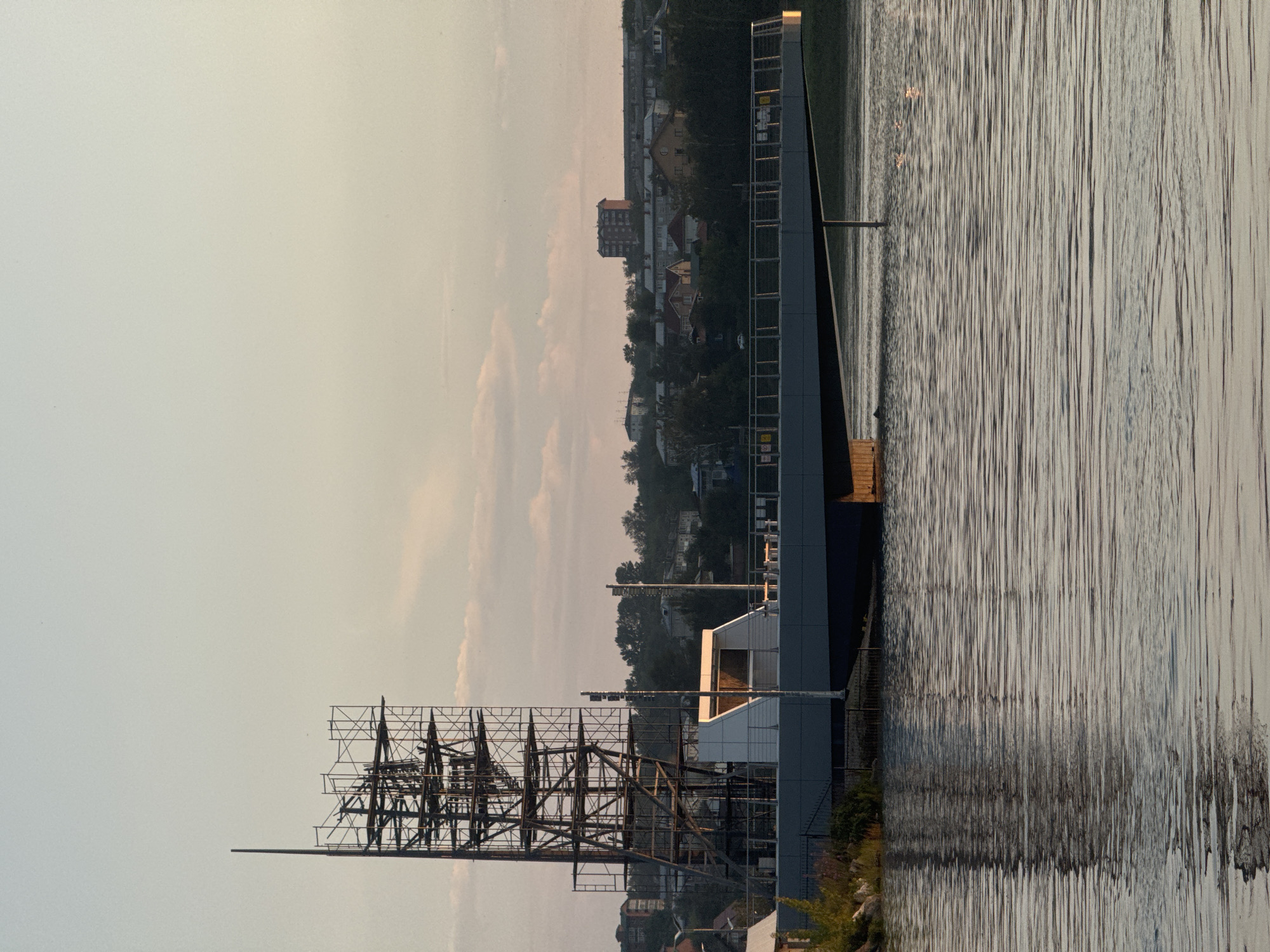 The image depicts a waterfront scene in Irkutsk, Russia, at what appears to be dusk or dawn. In the foreground, a body of water with gentle ripples reflects the sky and surrounding structures. The water is calm with subtle light glinting off its surface.
Dominating the mid-ground is a modern, angular structure extending over the water, possibly a bridge or a pier. This structure is characterized by its sleek, grey, horizontal lines and a prominent railing with vertical supports. To the left of this structure, a building with white, geometric walls and angled roofs is visible, suggesting a contemporary architectural design.
Rising behind these structures, and partially obscuring the cityscape, is an extensive network of scaffolding. This scaffolding is a complex, multi-tiered metal framework, indicative of ongoing construction or renovation. It is tall and imposing, creating a stark contrast with the more finished elements of the scene.
In the background, the city of Irkutsk unfolds. A hillside is densely populated with various buildings, ranging from smaller, residential houses with pitched roofs to taller, apartment blocks. Green trees and foliage are interspersed throughout the urban landscape, softening its appearance. The sky is a pale, muted gradient of blues and pinks, with soft, wispy clouds, further suggesting the twilight hour. No people are clearly visible in the image, contributing to a sense of quietude or recent activity.