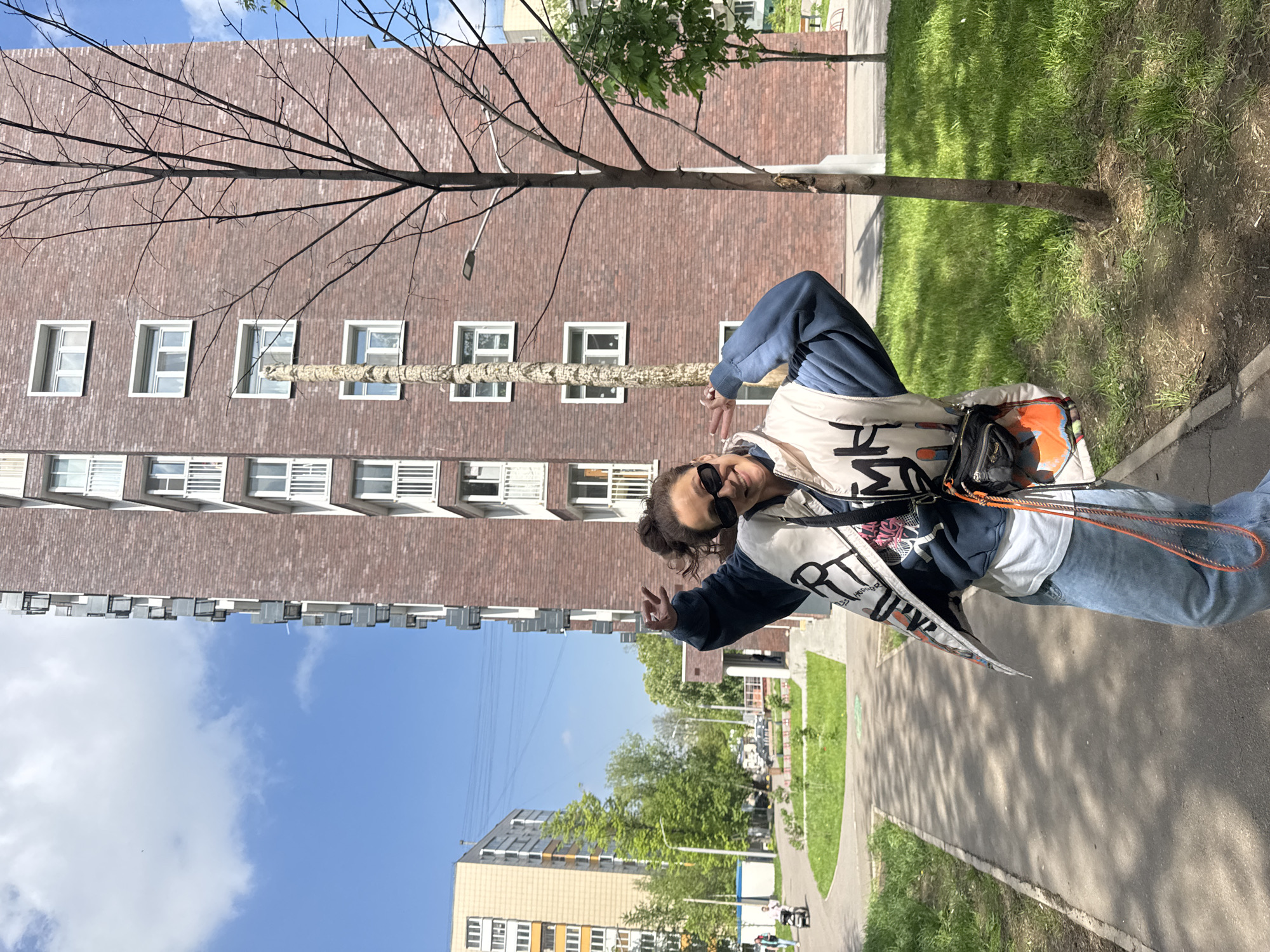 The image captures a person posing outdoors, with urban elements in the background. The individual wears sunglasses, jeans, a jacket, and a shoulder bag. They are making a hand gesture. The background includes apartment buildings, trees, a sidewalk, and patches of grass. The sky is blue with some clouds. Based on architectural style and general environment, the location appears to be in Moscow, Russia.