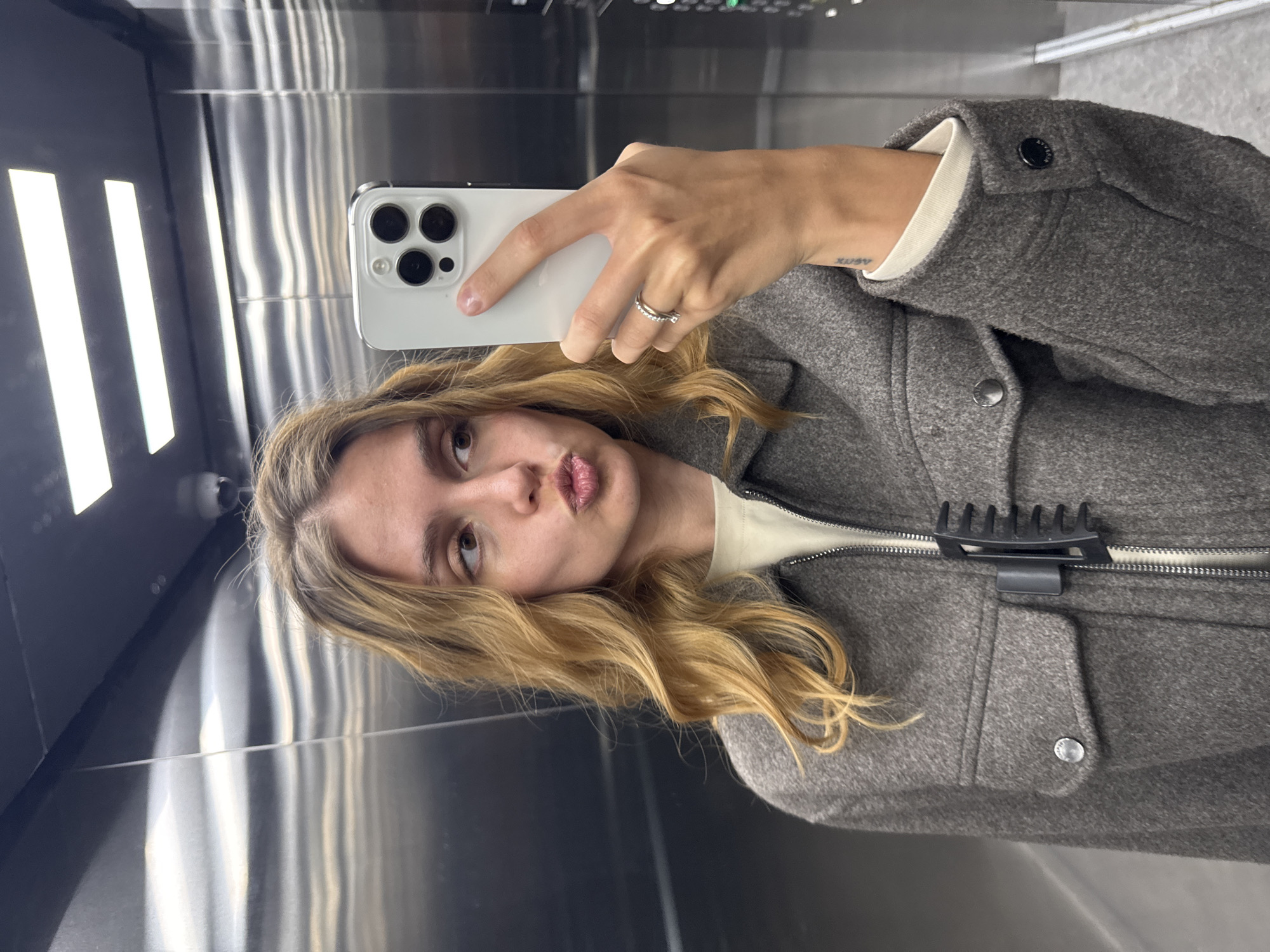 A woman is taking a selfie in an elevator. She is holding a white smartphone in her right hand. She has long blonde wavy hair and is wearing a grey wool coat over a white shirt. She is making a kissing face. The elevator has mirrored walls with vertical LED lights. The location is Unknown, Unknown.
