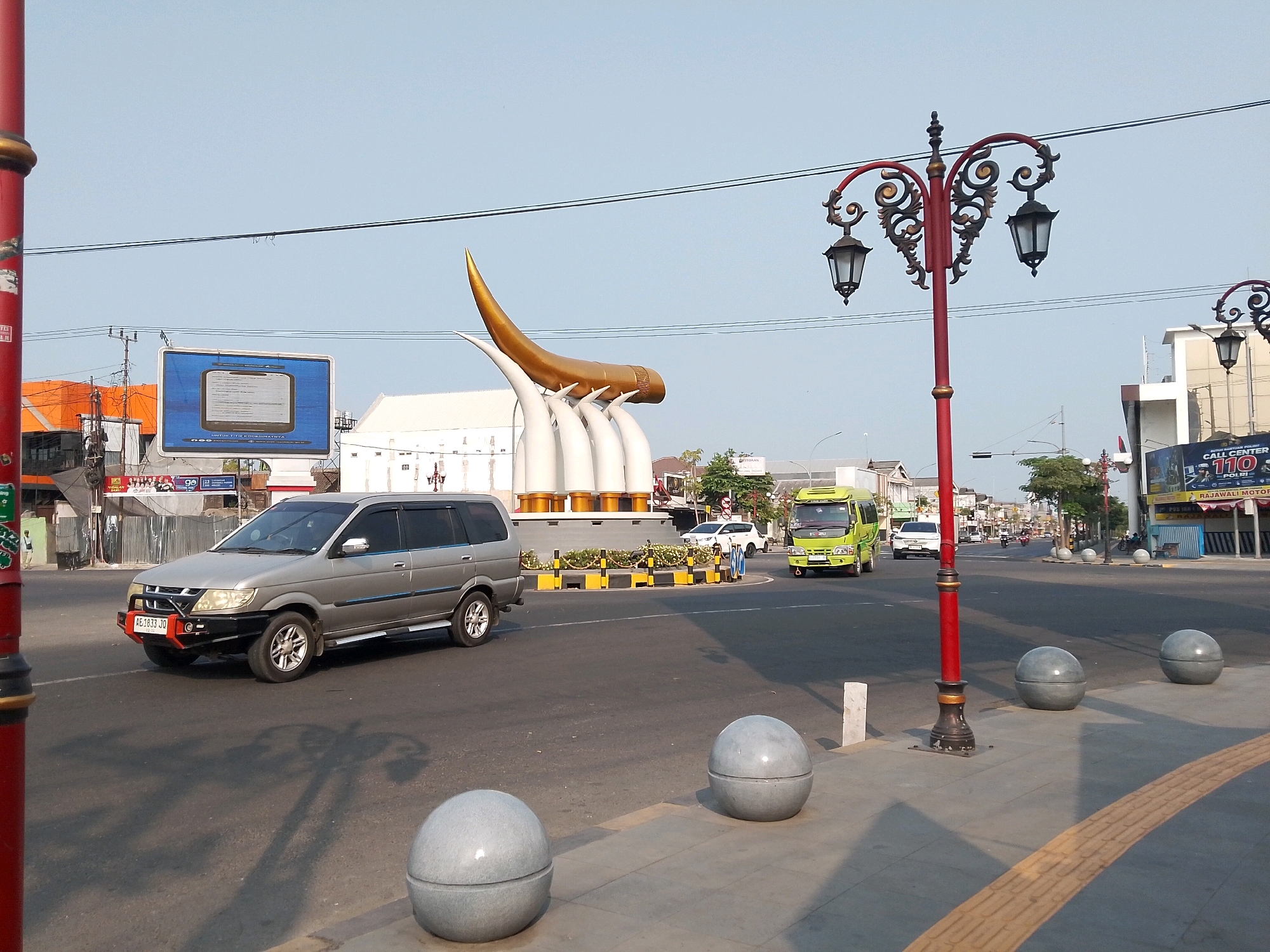 The image captures a street scene in the city of Ngawi, Indonesia, characterized by a bright, clear sky and warm sunlight casting long shadows. In the foreground, a grey SUV with a red and black bullbar is parked on the right side of a paved road. The SUV has license plate "AE 1833 JQ". To the right of the SUV, a sidewalk is visible with three large, spherical concrete bollards. The sidewalk features a textured yellow tactile paving strip.
Dominating the mid-ground is a prominent golden horn sculpture, part of a larger monument, supported by white pillars. This monument is situated at a junction or roundabout, bordered by yellow and black striped barriers. A decorative red lamppost with two black lanterns stands to the right of the monument.
Several vehicles are in motion on the road. A bright yellow van is driving past the monument, followed by a white car. Further down the road, more vehicles and a few pedestrians can be seen. Buildings line the street, including a structure with "CALL CENTER 110 POLRI" signage and another with an orange facade. The overall environment suggests a bustling urban area during the daytime.