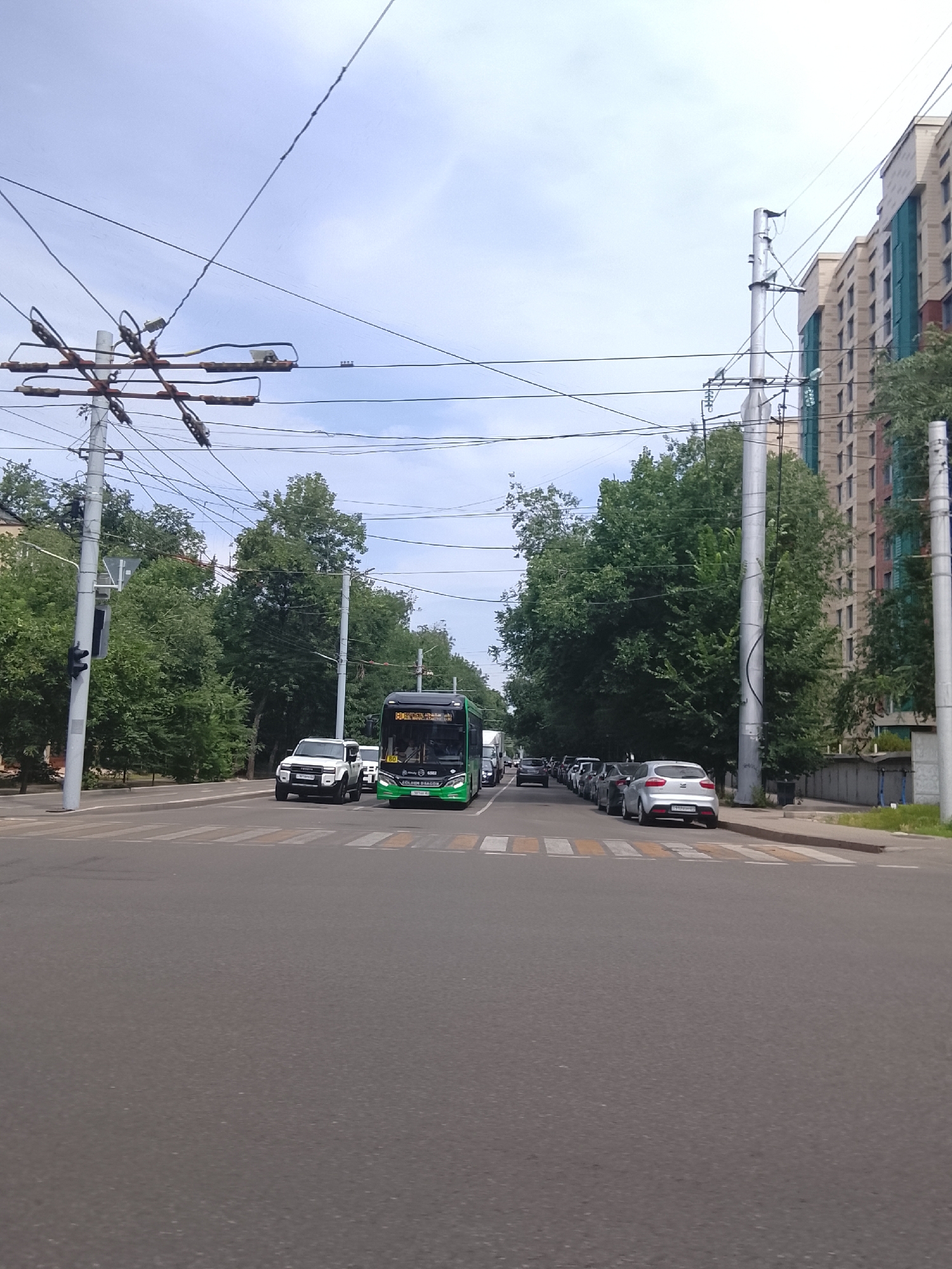 The media file captures a daytime street scene in Almaty, Kazakhstan. The image is dominated by a busy intersection with a pedestrian crossing in the foreground. Several vehicles are present, including a bright green bus, a white SUV, and a line of parked cars. Overhead, a complex network of power lines and utility poles crisscrosses the sky, indicating an urban environment with overhead electrical infrastructure. Trees line the street, providing a natural element within the city landscape. In the background, modern multi-story buildings rise, suggesting a developed area. The sky is partly cloudy, hinting at moderate weather conditions. The time appears to be daytime, given the lighting. No people are clearly visible in the scene. Text is visible on the green bus, including the number "80" and some Cyrillic characters that are difficult to fully decipher. License plates on some of the vehicles also display text. The overall impression is that of a functional urban setting during a typical day.