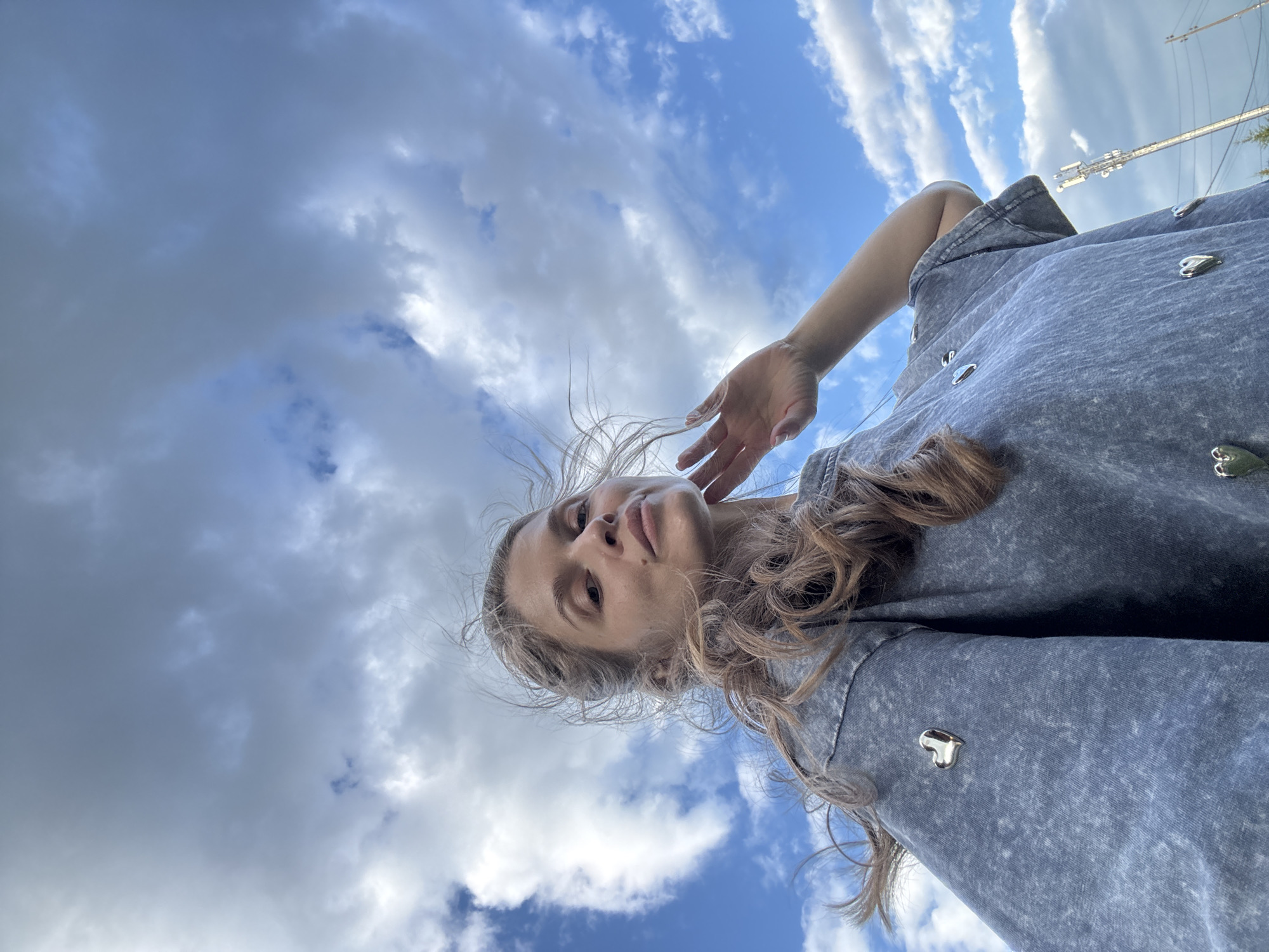 In Minusinsk, Russia, a woman with long, wavy brown hair, partially obscuring her face, poses for a selfie against a backdrop of a blue sky filled with fluffy white clouds. She is wearing a grey, acid-wash t-shirt adorned with multiple heart-shaped metallic embellishments. One hand is raised, touching her chin, while her hair blows in the wind. The image is taken from a low angle, emphasizing the sky. In the background, a cell tower and some distant greenery are visible.