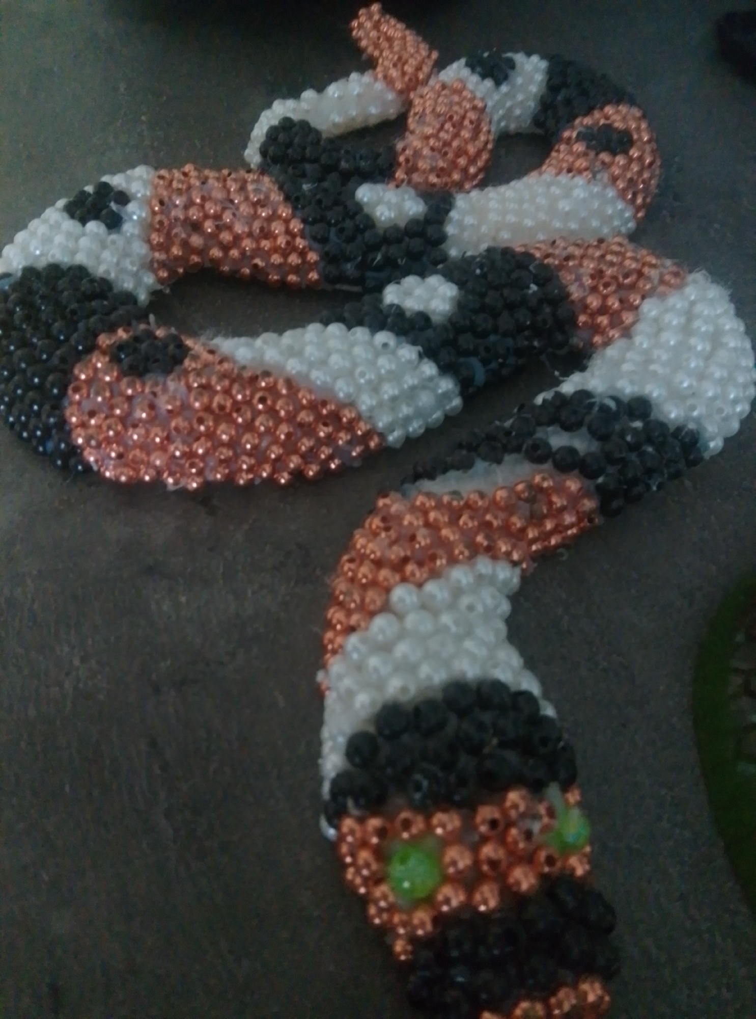 A beaded snake sculpture is arranged in a coiled position on a dark, textured surface. The sculpture is made of small, round beads in alternating sections of white, black, and copper. The head of the snake features two green beads for eyes and a black beaded mouth. The background is dark and indistinct, with a hint of green on the right side of the frame. The lighting is dim, casting shadows that obscure fine details of the environment.