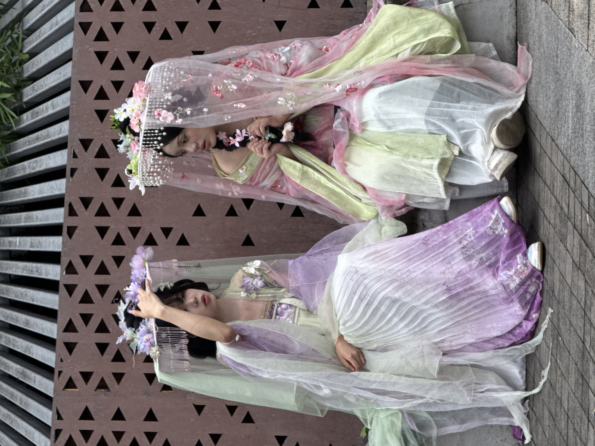 Two young women, dressed in traditional Chinese attire, are seated on a ledge outdoors. The woman on the left wears a layered garment in white and purple, with sheer white and pale green fabric draped around her. She has a headpiece adorned with flowers and pearl-like strands. Her right hand is raised near her face. The woman on the right is dressed in a pink and pale green outfit, also with a floral headpiece and sheer veil. She is holding a strand of flowers and her braid. Both women have their hair styled in dark buns. They are both wearing casual white sneakers. Behind them is a decorative brown wall with a triangular cutout pattern, and above that, a slatted structure. The ground in front of them is paved with rectangular stones. The scene appears to be in an urban outdoor setting, possibly Yuzhong District, China, given the architectural style and cultural context.