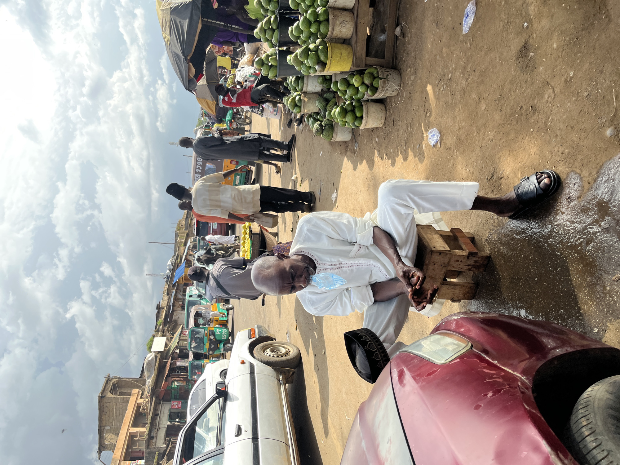 The image depicts a bustling outdoor market scene, likely in Rafin Par, Nigeria, under a cloudy sky. In the foreground, a man dressed in a white kaftan and pants sits on a small wooden stool next to a red car. He is holding a clear plastic bag filled with water to his mouth. A black skullcap rests on the ground near his feet. The ground is a mixture of dirt and wet patches, suggesting recent rain.
The market is lined with various stalls and vendors. To the left, several vehicles are parked, including a white car and multiple auto-rickshaws. Further into the market, a collection of fruit stalls are visible, laden with green avocados and other produce. Numerous people are seen walking and interacting within the market. In the background, buildings with weathered facades and stalls with awnings indicate the commercial nature of the area. The overall atmosphere is dynamic and active, characteristic of a busy marketplace.