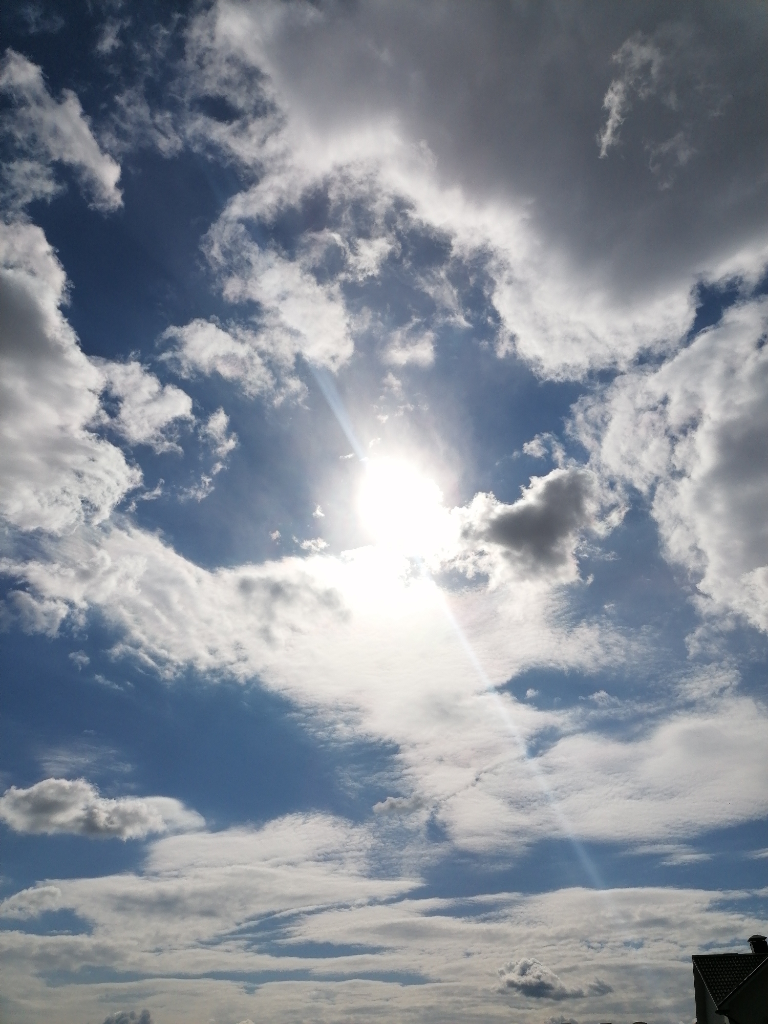 This media file captures a dynamic sky scene dominated by bright sunlight and various cloud formations. The central focus is the sun, a brilliant white orb, positioned in the upper middle of the frame. It shines intensely, partially obscured by clouds, with strong lens flares and distinct crepuscular rays radiating downwards and to the left through breaks in the cloud cover.
The sky is a vibrant blue in the clearer areas, contrasting with a diverse array of clouds. There are fluffy, bright white cumulus clouds scattered throughout, particularly on the left side, and broader, more diffuse cloud masses. Some clouds, especially towards the right and bottom, appear thicker and have darker grey undersides, suggesting varying levels of density and light absorption. Wispy, high-level clouds are also visible, adding texture to the expanse.
In the bottom right corner, a small portion of a building's dark-tiled roof and a chimney is visible, indicating the photograph was taken from a ground-level perspective, looking upwards.
The scene depicts a bright, sunny daytime, likely mid-day, characterized by a mix of sunshine and cloud cover, creating a visually engaging interplay of light and shadow in the sky. There is no visible text in the image.
