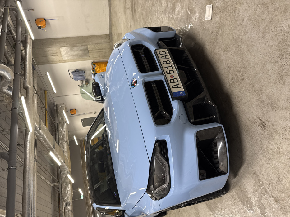 A light blue BMW M series coupe is parked indoors on a concrete floor. The vehicle features a black roof, black kidney grilles, dark-tinted headlights, and carbon fiber front bumper and air intake accents. A Slovakian license plate, 'SK AB•518 AG', is attached to the front bumper. The driver's side door is open, with the window partially lowered. Water residue is visible on the front bumper and a small puddle is present on the floor to the right of the vehicle. The interior space appears to be a car detailing or service bay, characterized by exposed concrete columns, light-colored walls, overhead fluorescent tube lighting, and silver ventilation ductwork. A yellow hose reel, a rack holding towels, and a yellow utility bin are visible in the background. No individuals are present. The specific location within Warsaw, Poland, cannot be determined from the visible evidence, as the license plate indicates Slovakia.