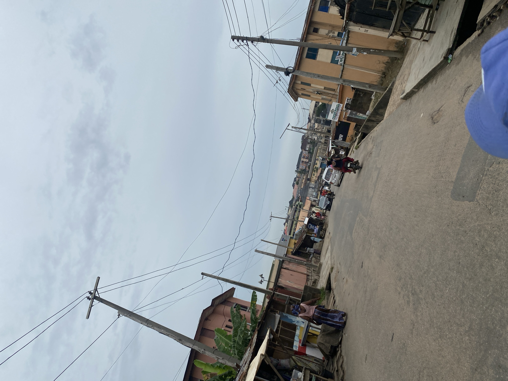 A street scene in Lagos, Nigeria. The road is paved but appears worn. Several buildings line the street, a mix of residential and commercial, mostly two-story structures with a few taller buildings in the distance. The architecture is simple and functional. Power lines crisscross overhead, supported by utility poles. A few people can be seen on the street, some on motorcycles, others are pedestrians or gathered near roadside businesses. There are cars and vans further up the road. The sky is overcast, suggesting it is likely the wet season. There is no specific landmark or business clearly visible that could definitively pinpoint the exact location.