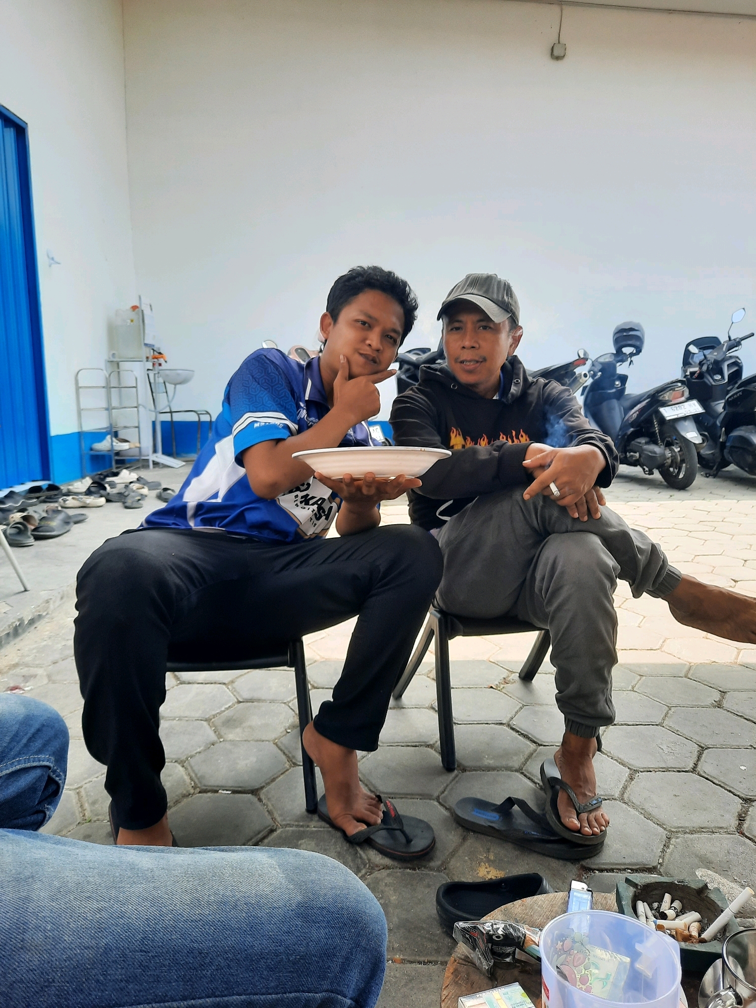 Two men are sitting on chairs outdoors. The man on the left is wearing a blue and white t-shirt and black pants. He is holding a white bowl in his left hand and has his right hand on his chin, looking at the camera. The man on the right is wearing a black hoodie with flame graphics and grey pants, a grey baseball cap, and a ring on his left hand. He is smoking a cigarette and looking to his left. There are several motorcycles parked in the background. The ground is paved with hexagonal tiles, and there are shoes and other items scattered on the ground. The scene appears to be in a public outdoor area, possibly near a building with a white wall and a blue door. The overall impression is a casual gathering.