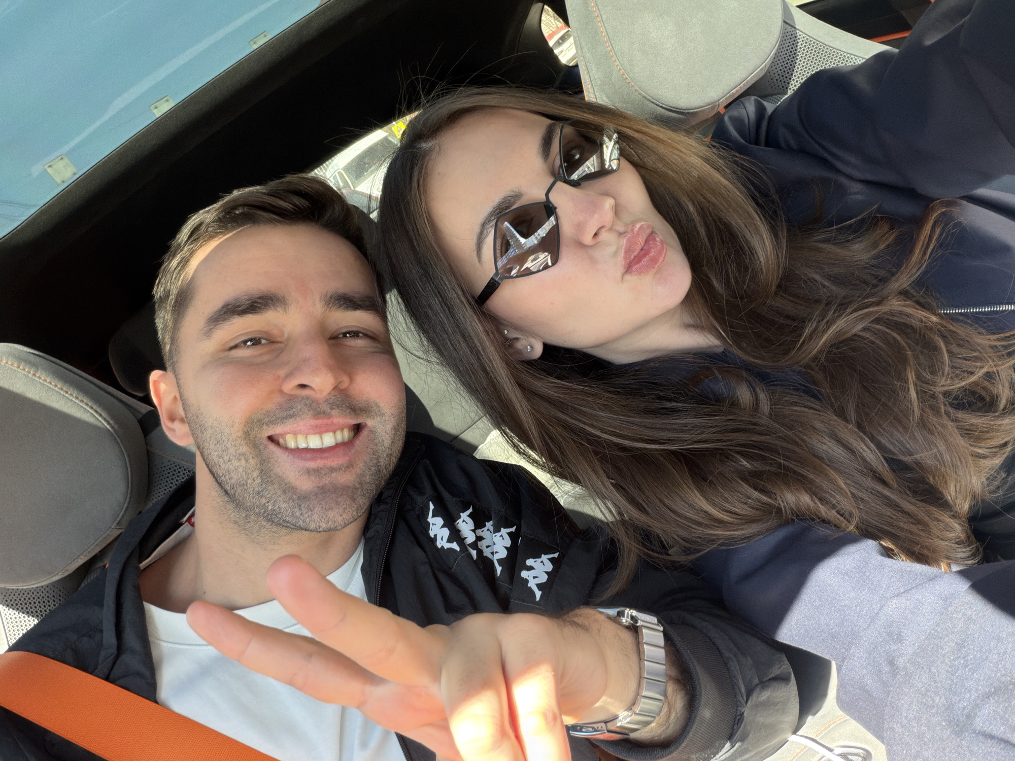 A bright, sunlit selfie taken inside a car captures two individuals, a man and a woman, during what appears to be a casual outing, possibly in Krasnoyarsk, Russia. The man, positioned on the left, has short dark hair and a light beard. He is smiling broadly, revealing his teeth, and forms a peace sign with his right hand, which is prominently featured. He wears a white t-shirt beneath a black jacket adorned with multiple white Kappa logos. An orange seatbelt crosses his chest, and a silver watch is visible on his left wrist.
The woman, to the right, has long, wavy dark brown hair. She wears stylish, angular dark sunglasses and a dark blue jacket. Her lips are pursed in a playful pout, highlighted with pink lipstick.
The setting is a car interior, suggested by the gray seats with orange stitching and a glimpse of a black roof lining. A clear blue sky is visible, reflected in what appears to be a sunroof or panoramic roof, indicating bright daytime conditions. Strong sunlight casts shadows on the subjects, highlighting the sunny weather. Both individuals convey a sense of happiness and playfulness through their expressions.