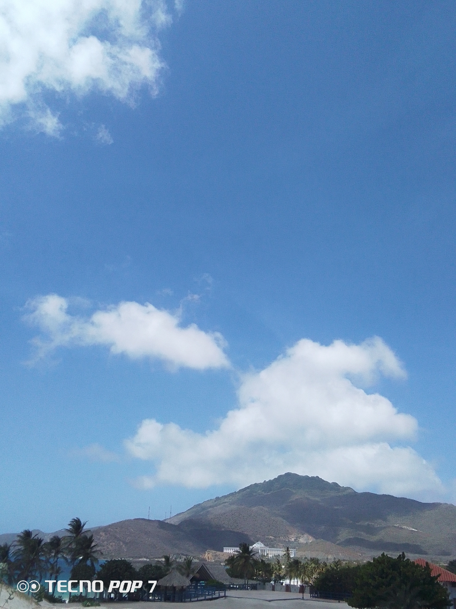 The image captures a daytime scene in Pedro González, Venezuela. The foreground features a view of the landscape, including a beach area with palm trees and thatched-roof structures. A large mountain dominates the midground, its slopes covered in vegetation and casting shadows. In the background, a large building or hotel is visible near the base of the mountain. The sky is predominantly blue with scattered white clouds. At the bottom left corner of the image, the watermark "TECNO POP 7" is present.