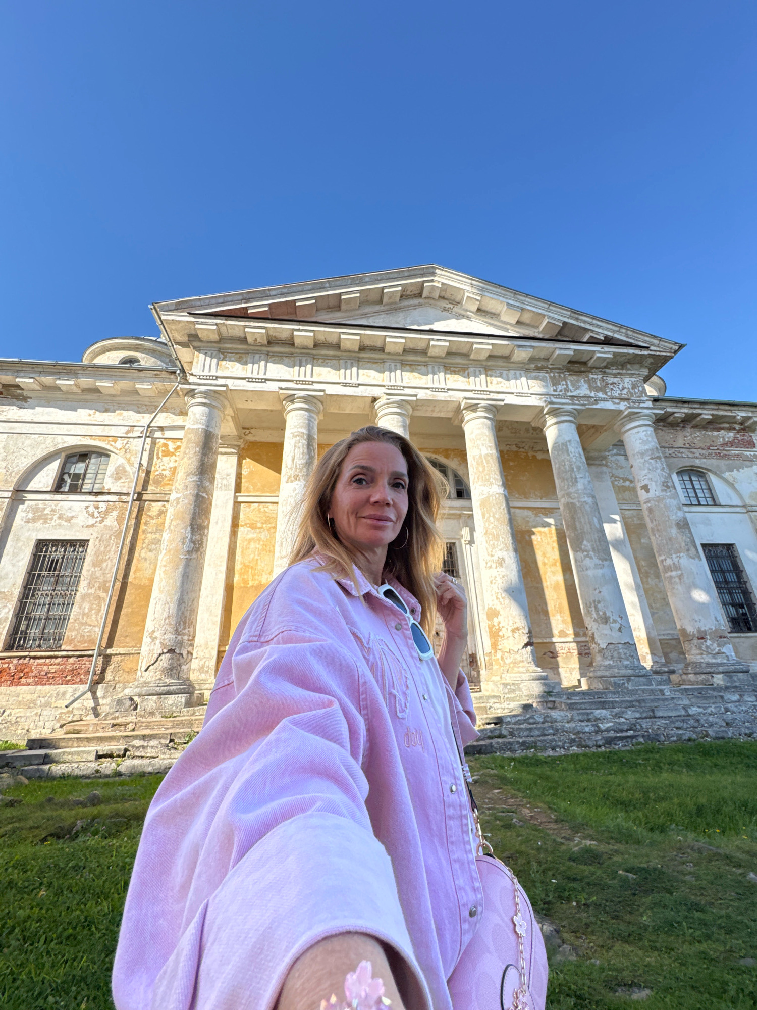 In the city of Torzhok, Russia, a blonde woman wearing a pink denim jacket stands in front of a large, weathered, neoclassical building with a colonnade. The building, appearing to be an abandoned estate or palace, has a pale yellow facade with peeling paint and visible signs of age. The woman is taking a selfie, holding the camera with her right hand, and her left hand is near her face. She is smiling at the camera, and her sunglasses are perched on her head. A light pink handbag with floral embellishments is slung over her shoulder. The sky above is a clear, bright blue, and the building is situated on a grassy incline. The sunlight casts shadows from the columns onto the building's facade.