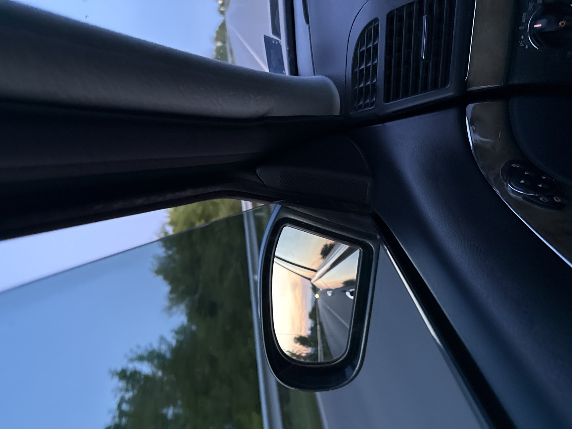 The image is an interior shot taken from inside a car, looking out towards the driver's side. The car's side mirror reflects a road stretching into the distance, with trees lining the side of the road. The sky in the reflection suggests either sunset or sunrise. The interior of the car is visible, including the air vents, window controls, and the door frame. The image does not provide any information about the location being in Mogila, Bulgaria or any people, subjects, actions, or interactions.