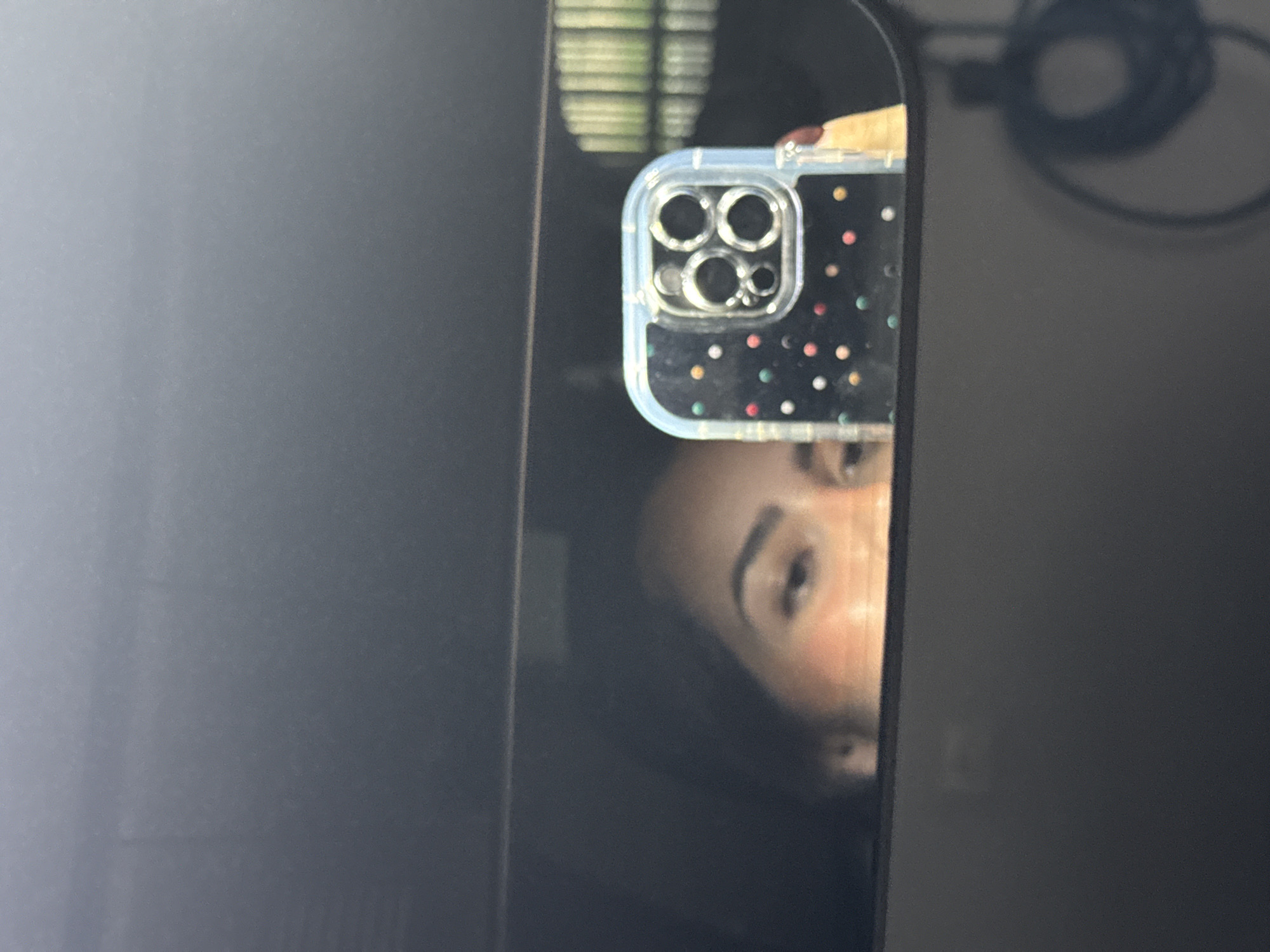 A person's partial face is reflected in a dark screen next to a phone, possibly an iPhone, with a clear case featuring colored polka dots. The phone's camera array is visible in the upper portion of the reflection. A glimpse of blinds is also visible in the screen's reflection. A black cable is in the background. The location of San Bernardino Tlaxcalancingo, Mexico, cannot be determined from the image.