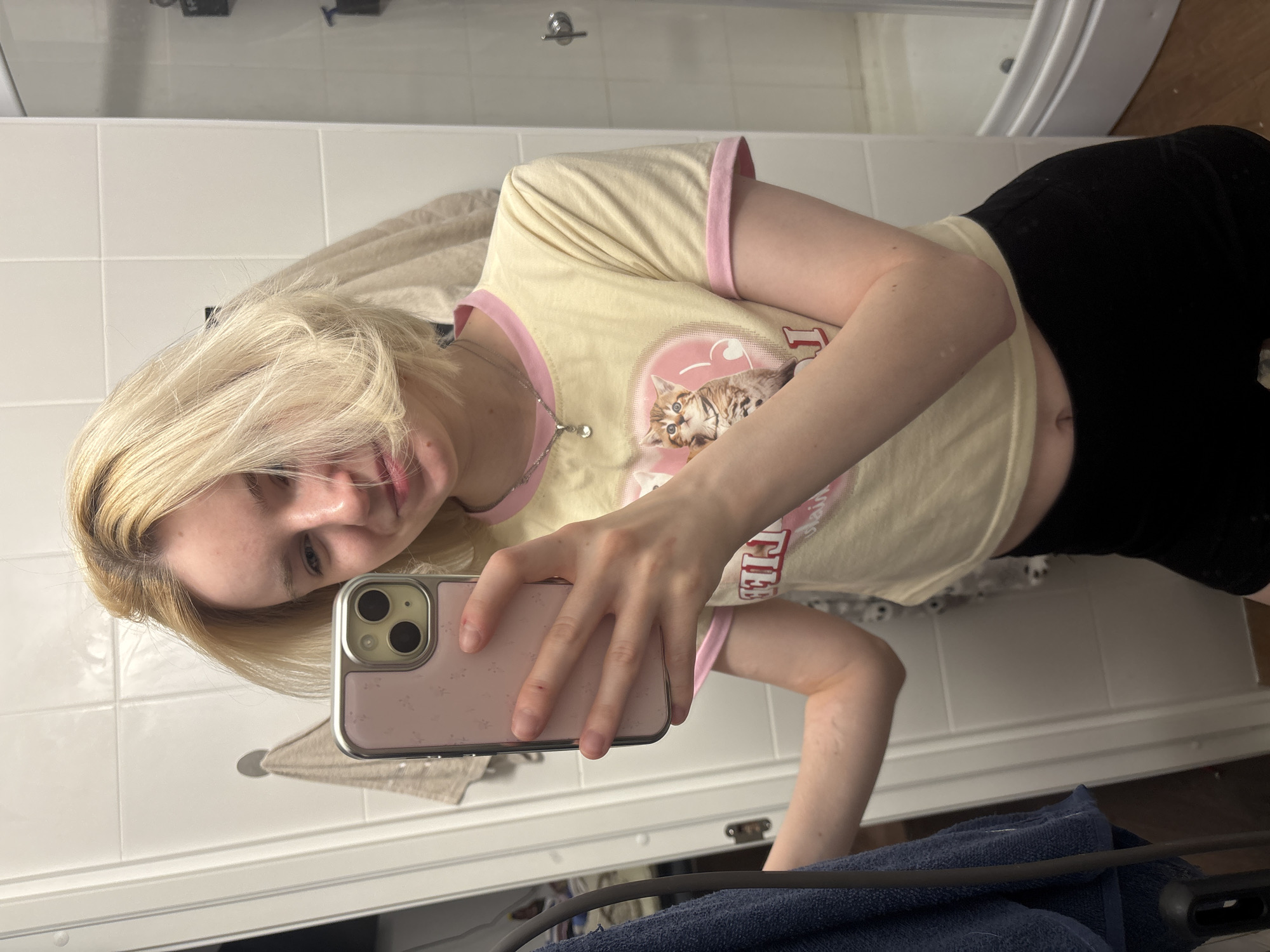 A young woman is taking a selfie in a bathroom. She has blonde hair and is wearing a light yellow t-shirt with pink trim and a graphic print of a cat, paired with black shorts. She is holding a pink iPhone in her right hand. The bathroom has white tiled walls and a white sink. The city of Vsevolozhsk, Russia cannot be determined from the image.