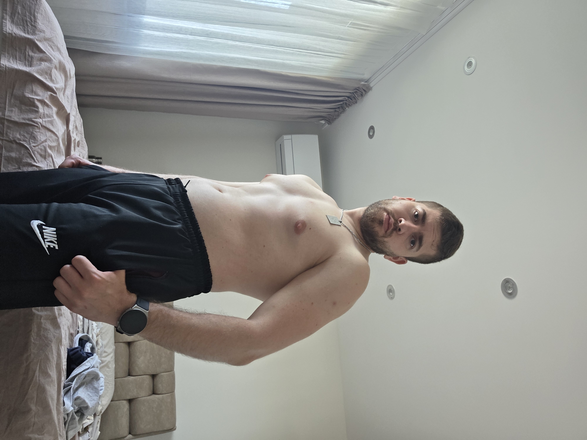 A fair-skinned, shirtless man with a beard and dark hair is wearing black Nike sweatpants and a smartwatch. He is standing indoors, next to a bed with beige linens. Curtains are visible behind him, and several recessed lighting fixtures are in the ceiling. An air conditioning unit is mounted on the wall. It is impossible to determine if this image was taken in Yagodovo, Bulgaria based on the given information.