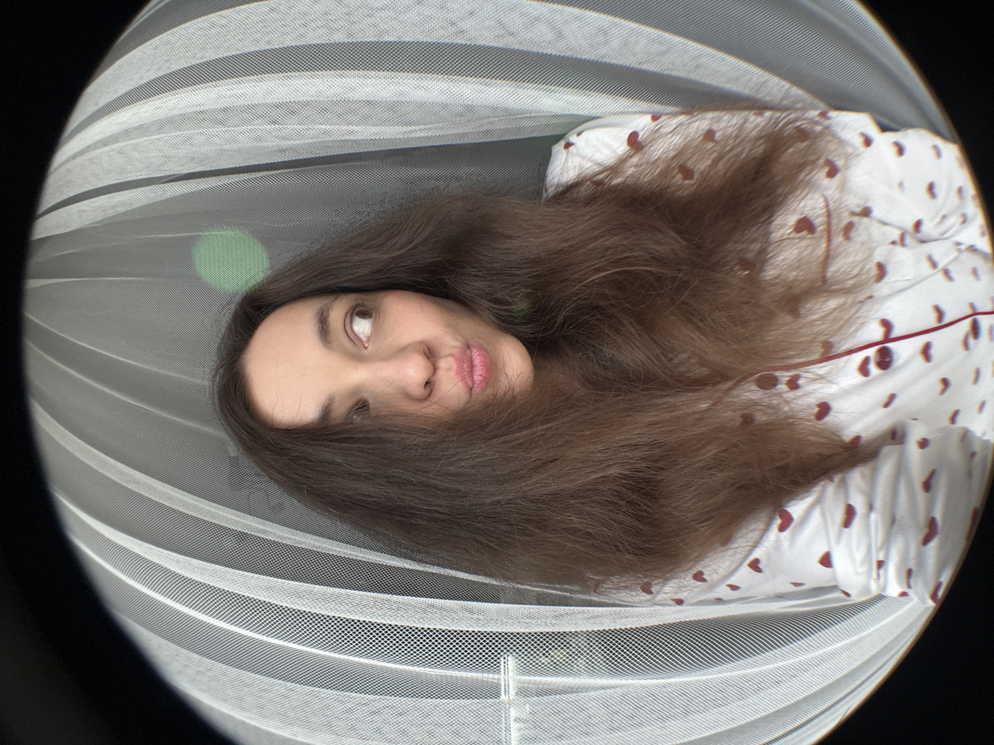 The image shows a woman lying down, possibly on a bed or similar surface, covered by a mosquito net or similar netting. She is wearing a white top with small red heart patterns. Her brown hair is spread around her head. The picture is taken with a wide-angle lens, creating a fisheye effect. A green reflection is seen on the net near her head. The location is Chaikovsky, Russia.