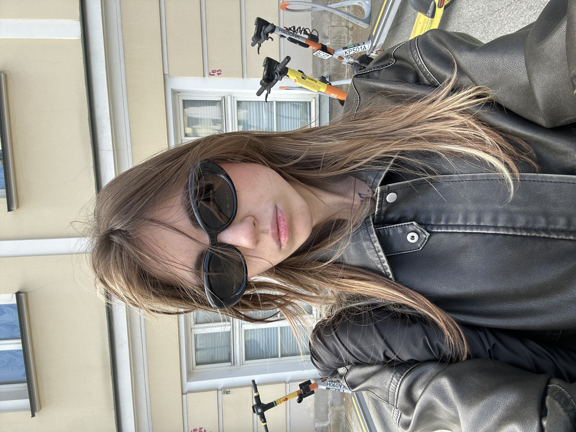 A young woman wearing sunglasses and a black leather jacket is taking a selfie outdoors. She has long, light brown hair that is partially covering her face. In the background, there are several rental electric scooters parked in front of a light yellow building with windows. The scene appears to be a city street. The woman is the primary subject, and the scooters and building are part of the environment. There are no visible indications of the specific city, nor any details to suggest it is Moscow, Russia.
