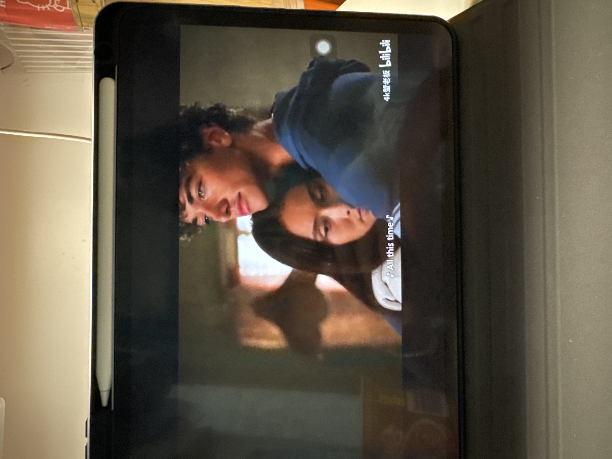 A tablet shows a still from a streaming video. Two people, a man and a woman, are the main focus. The man is positioned behind the woman, possibly hugging her. The video is on the Bilibili platform, indicated by the 4K label in Chinese. There is a music symbol next to "All this time." The tablet has an Apple Pencil attached. The scene is Nan'an District, China.
