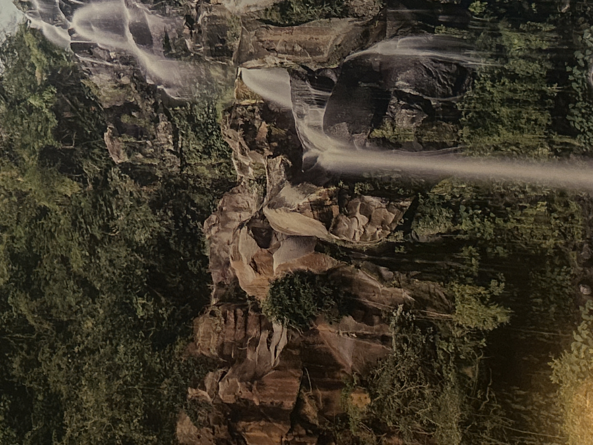 The image shows a waterfall cascading over rocky cliffs and lush greenery. The rocks are a mix of dark and light brown hues. The water is flowing rapidly, creating a white, blurred effect as it falls. The surrounding vegetation consists of dense green foliage. There is no evidence of any specific location in Plovdiv, Bulgaria, people, subjects, objects, actions, or interactions in the image. The environment is primarily natural with rocks, water, and plants dominating the scene.