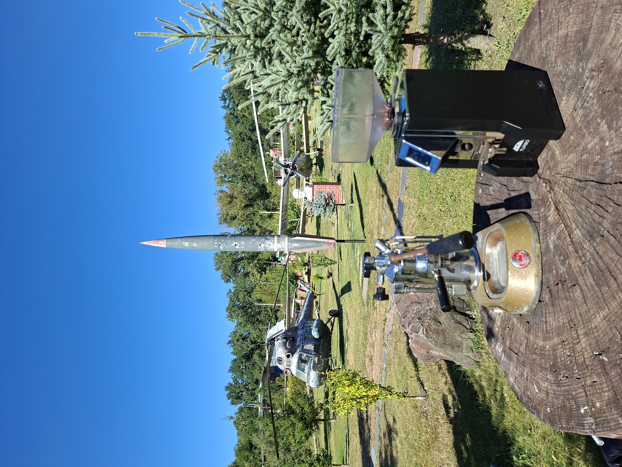 In Malcoci, Moldova, an outdoor scene unfolds with a clear blue sky overhead. In the foreground, resting on a weathered wooden stump, are two coffee-making devices: a vintage silver espresso machine with a worn brass base and black handles, and a modern black coffee grinder with a digital display and a clear plastic hopper containing coffee beans. Behind these items, the landscape transitions to a grassy park-like setting dotted with trees. Dominating the middle ground are several decommissioned military aircraft, including a helicopter with its rotor blades extended and a missile standing upright. Further in the background, more trees and a building are visible, suggesting the location is a museum or a historical park. The overall impression is one of an unusual juxtaposition of vintage and modern technology against a backdrop of military history and natural surroundings.