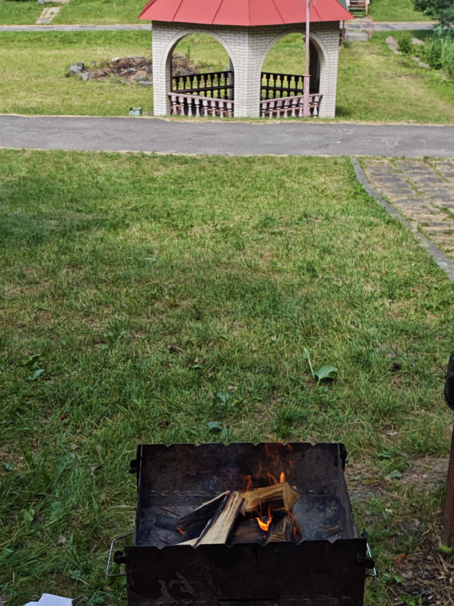 The image captures a scene in Sosny, Ukraine, with a focus on a barbecue grill in the foreground. The grill, a dark metal brazier, contains burning wood, with flames licking at the logs. The surrounding area is covered in green grass, suggesting an outdoor setting.
In the background, a small, open-sided structure with a red roof stands prominently. It's built of light-colored bricks and features arched openings with decorative wooden railings. Beyond the structure, more grassy slopes and a paved pathway are visible.
The overall impression is one of a casual outdoor gathering or a preparation for a barbecue. The natural lighting suggests it is daytime, likely with mild weather. The setting appears to be a park or recreational area. There is no visible text in the image.