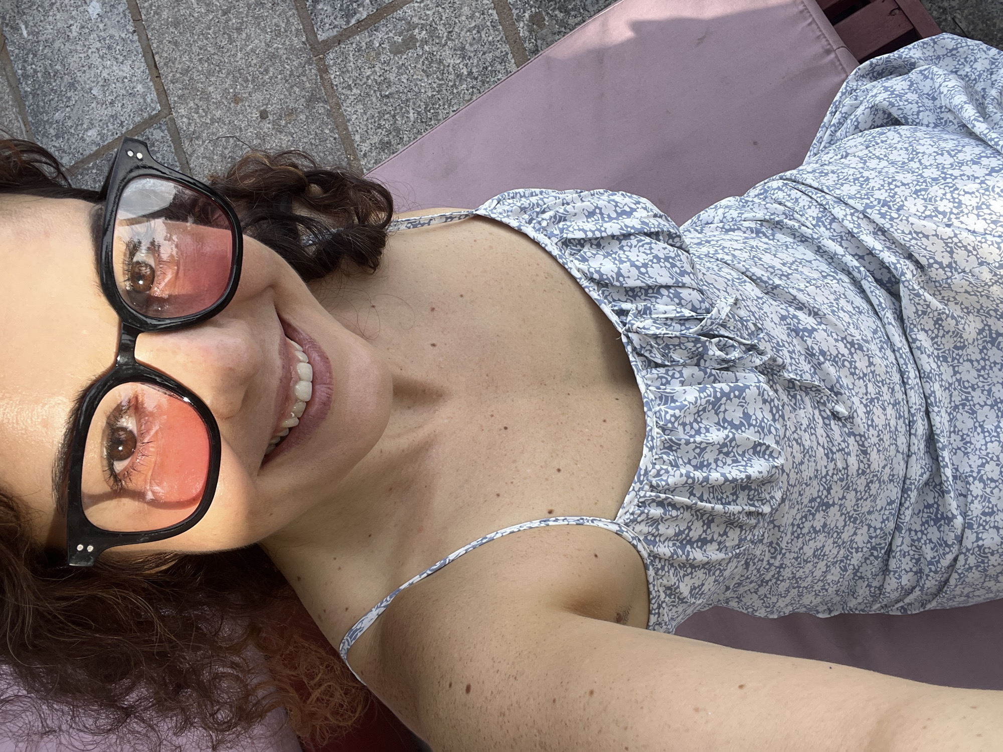 A woman with curly brown hair is seen from the chest up, wearing black-rimmed sunglasses with pink lenses and a light blue floral print dress. She is smiling, with her teeth showing. Her left arm and shoulder are visible, and she has freckles on her skin. She appears to be sitting on a pink lounge chair. The background shows a stone patio and what looks like red and dark brown wood, possibly part of the lounge chair's frame. The scene is outdoors and appears to be during the daytime, with sunlight casting shadows. There is no visual information in the image to suggest the location is Mũi Né, Vietnam.