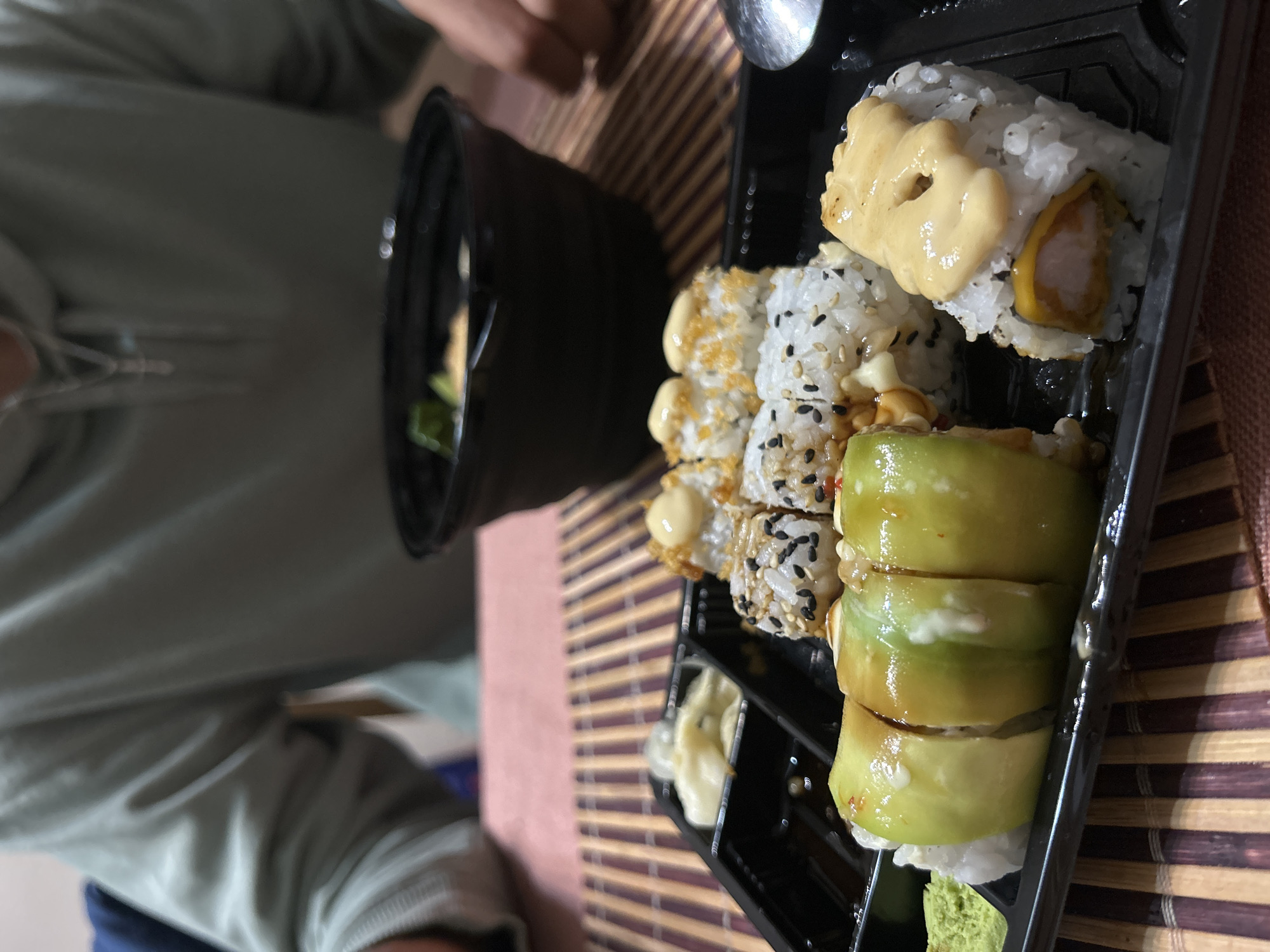 The image shows a black plastic container of sushi on a brown and beige bamboo mat. The sushi includes rolls with avocado, sesame seeds, and sauce. A small container of what appears to be salad sits to the left of the sushi. A person wearing a light green hooded sweatshirt is visible partially in the background. A spoon is near the upper right corner of the sushi. The location is Unknown, Unknown.