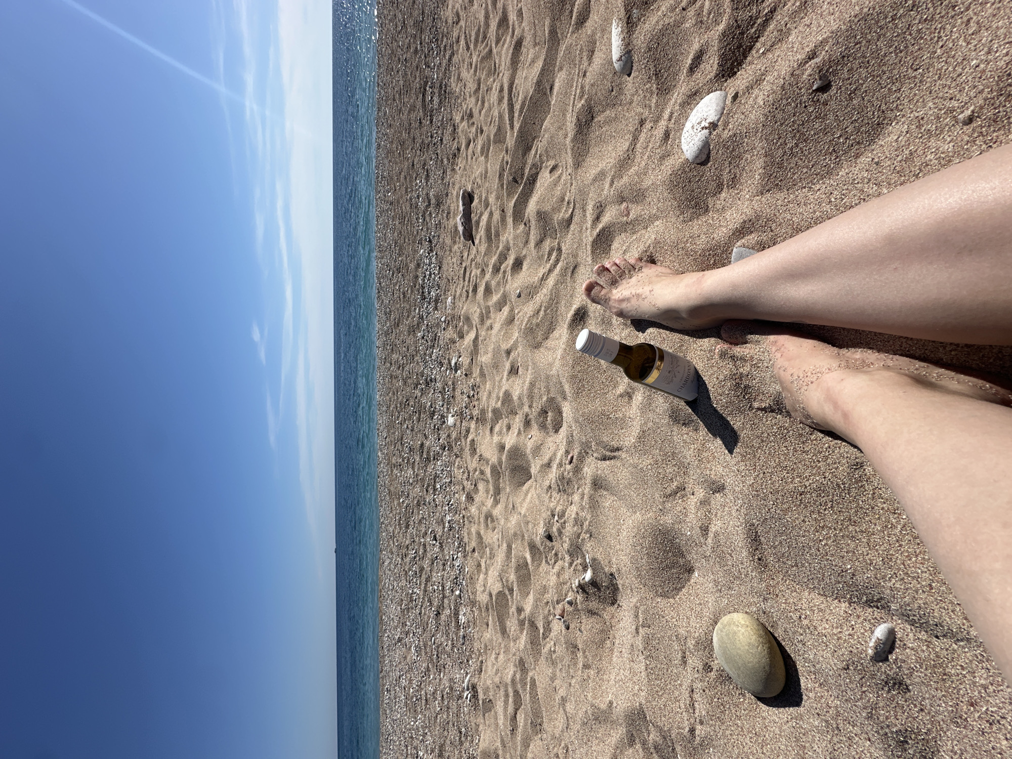 A person's legs and feet are resting on a sandy beach next to a small bottle of wine. The beach is located at Mirište, Montenegro. The sand is golden brown and contains scattered pebbles and stones. Beyond the beach, a calm, turquoise sea stretches toward the horizon under a clear, blue sky.