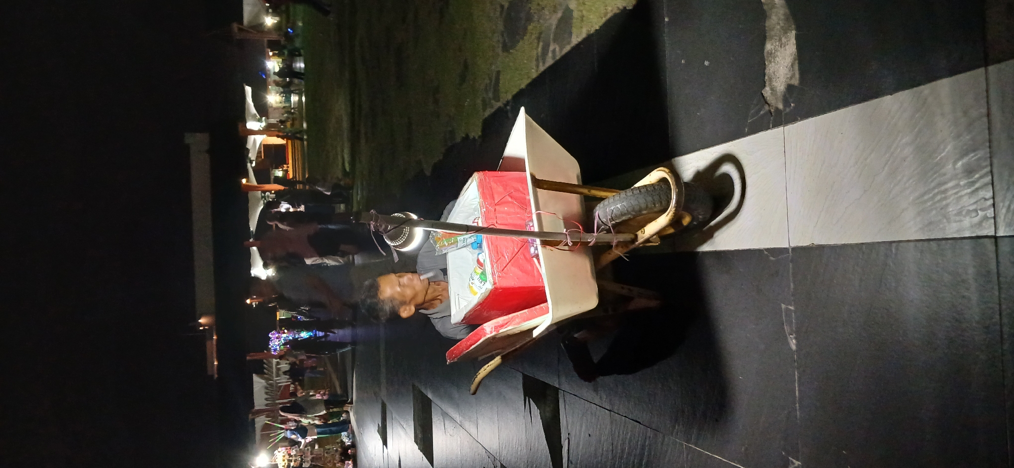 In Tanjung Balai Karimun, Indonesia, a man sits in a wheelbarrow at night. The wheelbarrow contains a red cooler box filled with bottled drinks. A lamp is attached to the wheelbarrow's handle, illuminating the man and the cooler. In the background, a crowd of people is visible, with stalls and lights suggesting a marketplace or event. The ground is paved with black and white tiles, and shadows stretch across the scene.