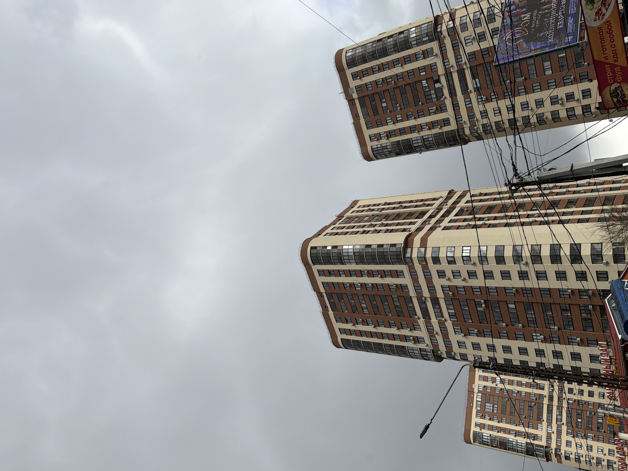 The image shows an upward view of three buildings against a cloudy gray sky. The buildings have a consistent architectural style, featuring light beige facades with horizontal rows of brown windows and brown accents. Utility wires are crisscrossing in front of the buildings and the sky. On the right side of the image, a street lamp, part of a banner, and additional signage are visible. Based on the context, the location appears to be in the city of совхоза "Воронежский", Russia.