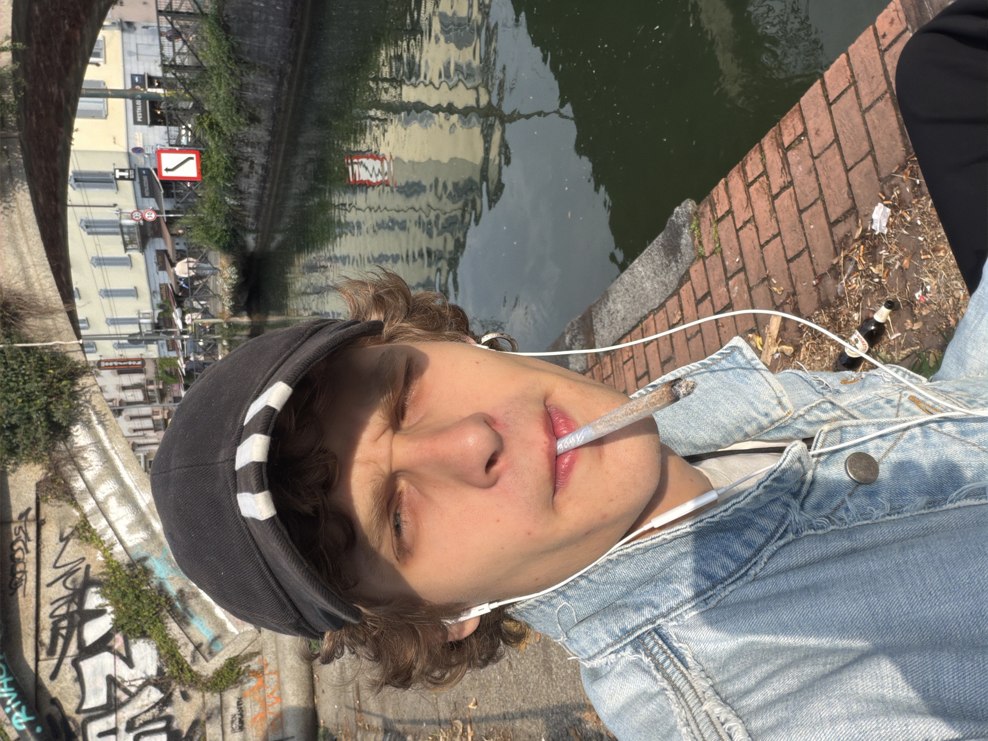 The image, rotated 90 degrees counter-clockwise, depicts a young person relaxing by an urban canal, consistent with the Navigli district in Milan, Italy. The central subject is a young individual with light brown, curly hair and fair skin, captured in a close-up. They are wearing a black cap with white stripes, a light blue denim jacket over a white t-shirt, and white wired earphones. A cigarette or joint is held between their lips, and their eyes are squinted, possibly due to bright sunlight or an expression of relaxation.
The immediate surroundings show a brick pathway next to dark green canal water. On the ground beside the path, there is visible litter, including numerous cigarette butts and an empty 'BIRRA MORETTI' beer bottle. To the left of the person, a concrete wall or structure is covered in green vines and graffiti, with stylized, illegible text.
Across the canal, reflections of buildings and the sky shimmer on the water's surface. On the far bank, multi-story buildings with light-colored facades are visible, along with road signs. One sign is a red square with a black upward-curving arrow, and another is a circular speed limit sign indicating "30". Partial text "POSTOLOF..." is also visible on a building's sign.
The scene is bathed in bright sunlight, suggesting a clear day, likely late morning or afternoon. The overall atmosphere is casual and relaxed, indicative of a moment of leisure in an urban environment.