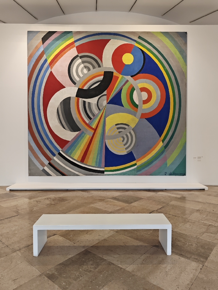 The image displays an interior view of an art gallery at the Centre Pompidou in Paris, France. A large, square abstract painting by Sonia Delaunay, identified by the signature "S. Delaunay" in the lower right, occupies the central portion of a plain white wall. The artwork, titled "Rhythm, Color, No. 700," features a complex arrangement of concentric circles, arcs, and radial segments in a vibrant palette of red, blue, yellow, green, and various shades of grey and black, along with rainbow spectrums. Below the painting, a low, elongated white barrier or plinth runs horizontally along the wall. In the foreground, a sleek, minimalist white bench with a rectangular shape is positioned centrally on a floor composed of large, light-toned stone tiles laid in a diagonal pattern. The ceiling, partially visible at the top, is white with subtle linear patterns. No individuals, specific actions, or interactions are present in the scene.
