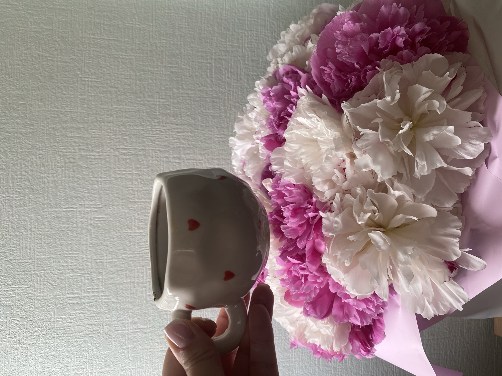 A close-up shot shows a hand holding a white mug with red hearts on it, positioned against a bouquet of white and pink peonies. The backdrop is a textured white wall. The image was likely taken indoors. No information indicates that this image was taken in the city of Sochi, Russia.