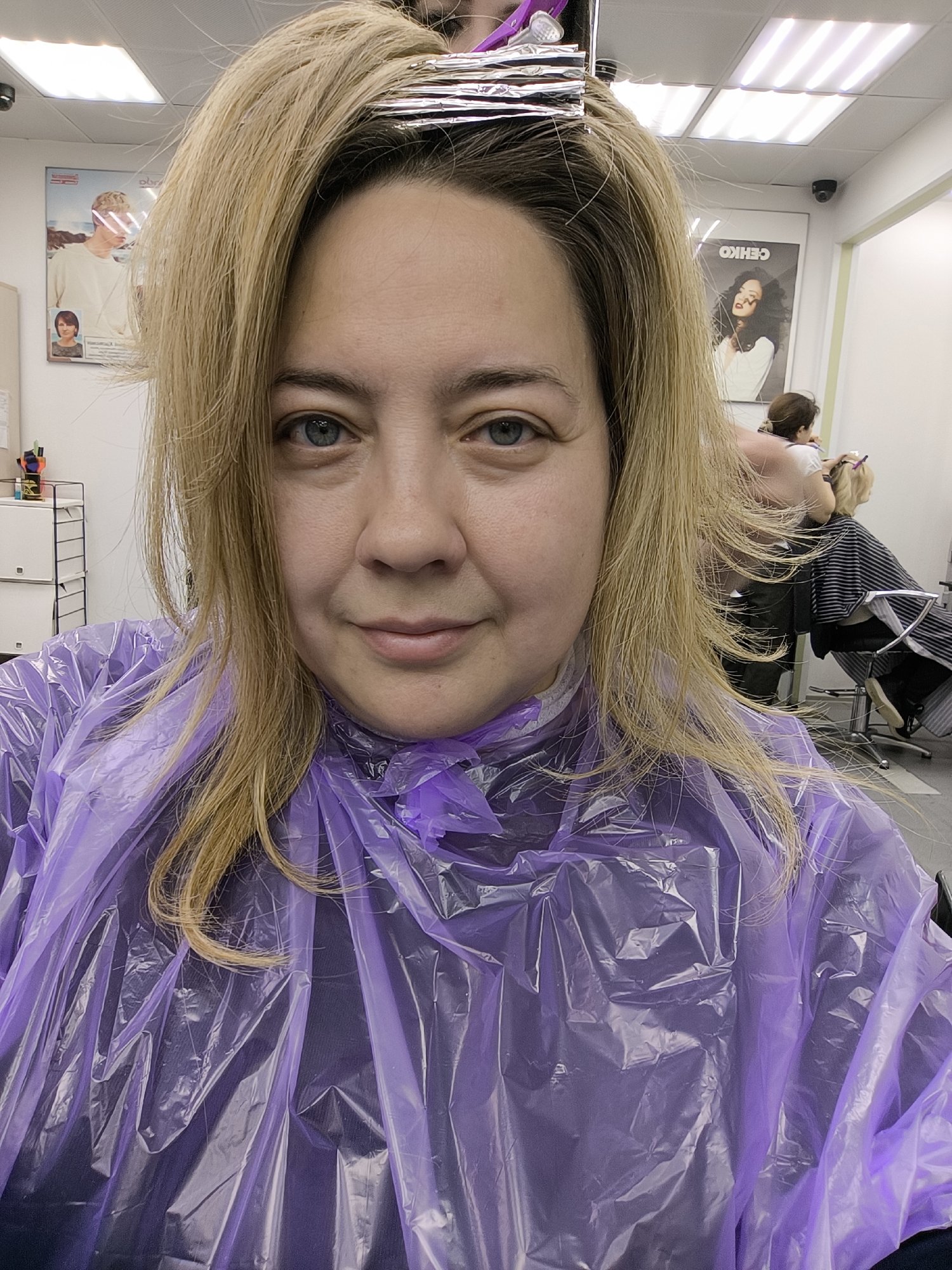 A woman with blonde hair is taking a selfie at a hair salon. She has foil in her hair for highlights and is wearing a purple plastic cape. In the background, another person is getting their hair styled. Posters of models with hairstyles are visible on the walls. The scene is taking place in Khimki, Russia, as indicated by the "CEHKO" poster, a brand commonly found in Russian hair salons.
