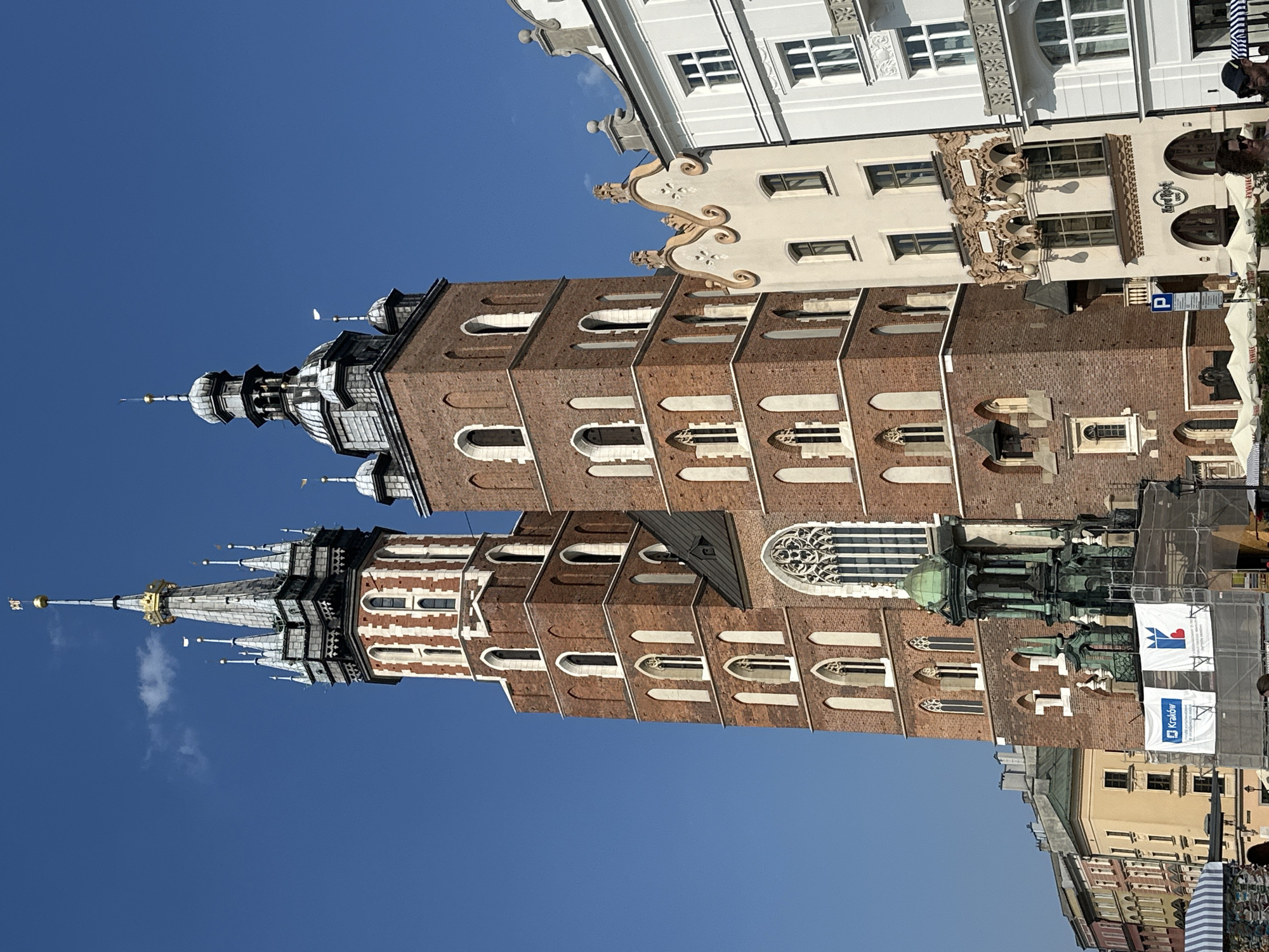 This is a photograph of St. Mary's Basilica in Krakow, Poland, taken from the Main Market Square. The basilica's iconic brick towers dominate the frame, with the taller, left tower featuring a gold-capped spire and the right tower having a more domed roof. The intricate gothic architecture of the church is visible, including arched windows and decorative stonework. To the right of the basilica, a row of colorful buildings with ornate facades lines the square. In the foreground, scaffolding and banners are visible around a monument in front of the church, suggesting ongoing restoration or an event. A few people are present in the lower part of the image, some of whom appear to be tourists. The sky is clear and blue, indicating a sunny day.