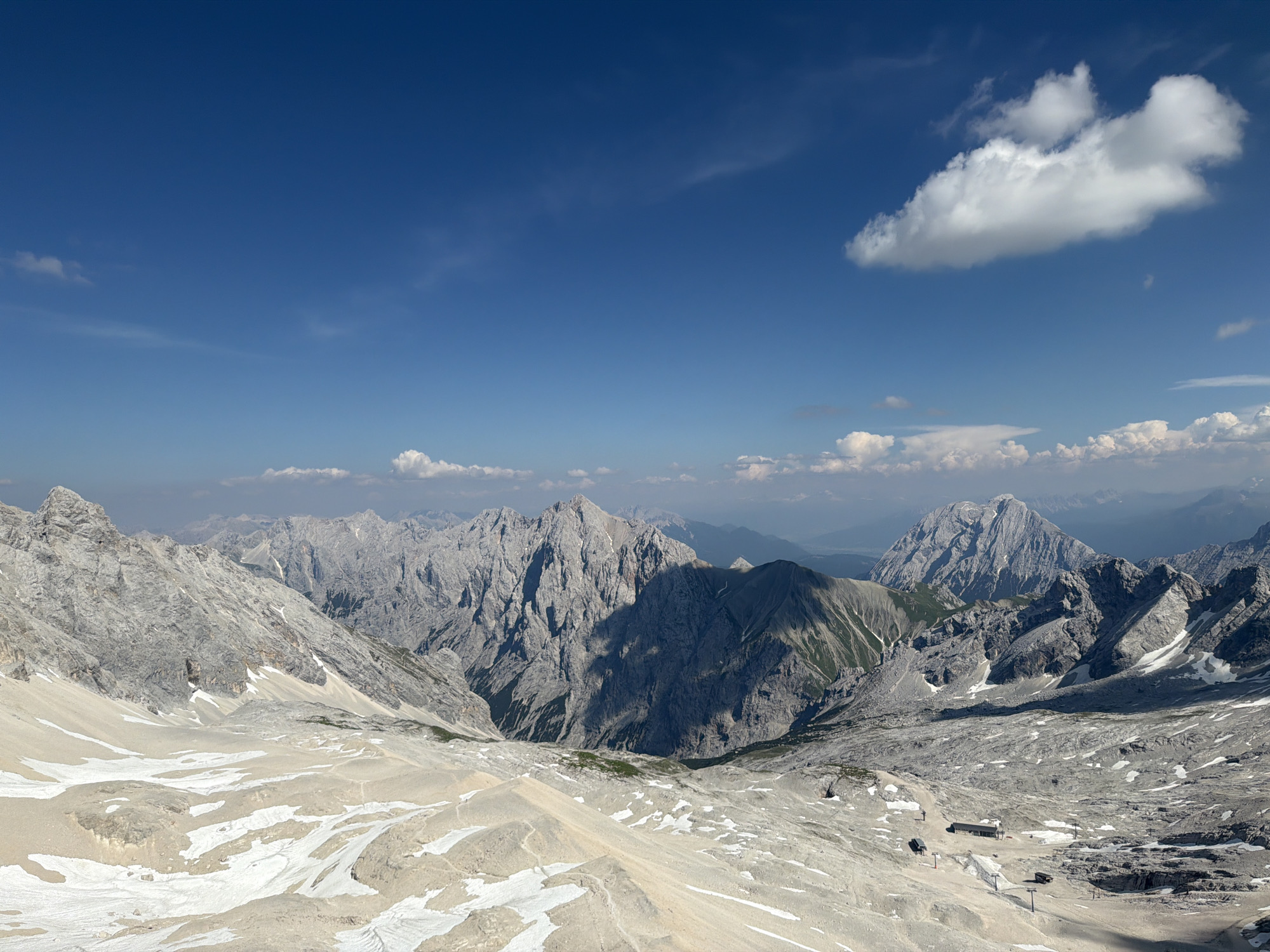 This is a high-angle, wide-shot landscape photograph taken from a mountainous vantage point. The scene is dominated by rugged, rocky mountains under a bright blue sky with scattered white clouds. The terrain in the foreground is a mix of scree and patches of snow, suggesting a high-altitude, possibly alpine environment.
In the middle ground, steep, gray, and jagged mountain peaks rise dramatically. Shadows are cast across these mountains, indicating the sun is relatively high in the sky, likely mid-day or early afternoon. Some of the mountain slopes show patches of green vegetation, contrasting with the predominantly rocky and snowy surfaces.
In the distance, a range of further mountains is visible, appearing softer and more hazy due to atmospheric perspective. The sky is a vibrant blue, with a few distinct white clouds, including a larger, more prominent cumulus cloud on the right side of the frame.
The most notable details are the vast, imposing mountain ranges and the stark contrast between the rocky textures, white snow, and blue sky. There are no visible people or specific activities taking place, lending a sense of natural grandeur and solitude to the scene. The location appears to be a remote, natural mountain area, possibly with some infrastructure like ski lifts visible in the lower-middle ground on the right. The overall impression is one of a clear, sunny day in a high mountain region.
No visible text.