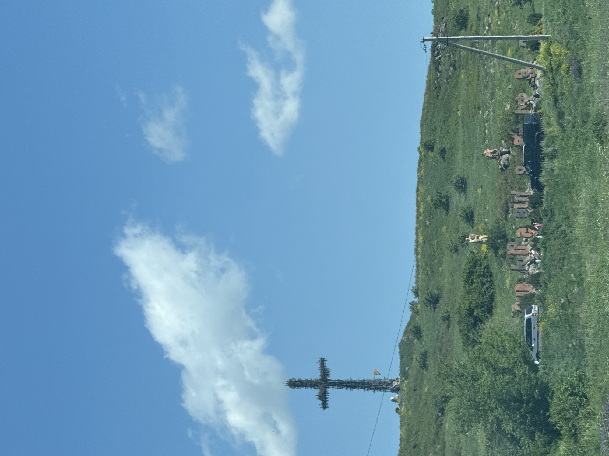 This image captures a panoramic view of the Saghmosavan Alphabet Monument and the Cross of the Fallen in Saghmosavan, Armenia. In the foreground, a grassy hill slopes upwards, dotted with green bushes and scattered yellow wildflowers. Several cars, including a silver minivan and a black SUV, are parked amidst the vegetation.
Further up the hill, large stone letters, forming Armenian script, are visible. These letters appear to be part of a monument, with some displaying weathered surfaces and graffiti. In the background, a towering metallic cross, known as the Cross of the Fallen, stands prominently against a clear blue sky with scattered white clouds. The cross is constructed from dark metal and has a rustic, unfinished appearance. A small flag, possibly red and white, is affixed to its base.
The environment suggests a sunny day with a gentle breeze, as evidenced by the white, puffy clouds. The overall scene conveys a sense of history and cultural significance, with the monuments serving as landmarks against the natural backdrop of the Armenian landscape.