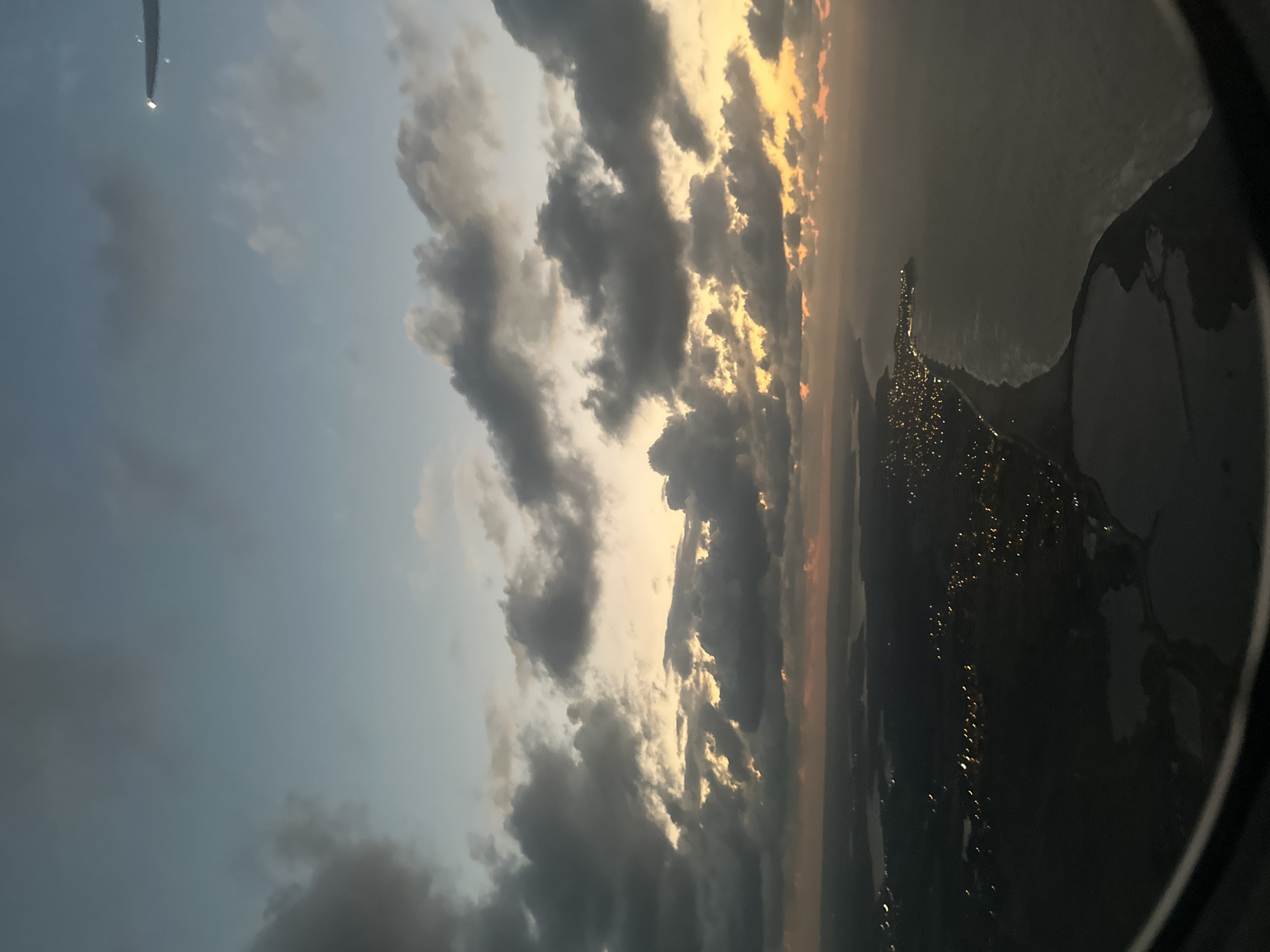 The image, taken from an airplane window, shows a sunset over a coastal city. The sky is filled with dramatic, dark clouds illuminated by the orange and yellow hues of the setting sun. Below, the city of Katunayake, Sri Lanka, is visible as a stretch of lights along the coastline. The ocean meets the shore, and there appear to be bodies of water within the city limits, possibly lagoons or marshlands. The edge of the airplane's wing is visible in the upper right corner of the frame.