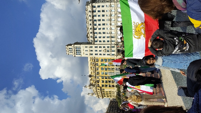 The image depicts a public gathering in Plaça de Catalunya, Barcelona, Spain, under a partially cloudy blue sky. In the background, two prominent buildings are visible: to the right, the Banco Sabadell building, a light-colored structure with a distinctive rectangular tower, and to the left, a large, ornate yellow building with a rounded corner.
In the mid-ground and foreground, a diverse group of people is assembled. Several individuals are holding flags; the most prominent flag, stretching across the lower right of the frame, is the historical Iranian flag, characterized by horizontal green, white, and red stripes with the golden "Lion and Sun" emblem in the center. Other smaller versions of this flag are also visible, along with what appears to be a Kurdish flag in the mid-ground.
In the immediate foreground, a young girl with glasses, wearing a light grey long-sleeve shirt and denim overalls, smiles towards the camera. Adjacent to her, a boy in a black hooded sweatshirt with a white and green graphic print stands with his back to the viewer, his hands placed on his head. Parts of other individuals are visible in the closest foreground, facing away from the camera. The ground is paved, indicating an urban square setting. The overall scene suggests a daytime public demonstration or rally.